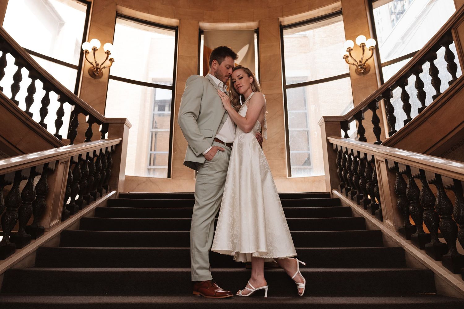 Bride and groom embracing on grand staircase in elegant venue, bride in tea-length white dress, groom in light suit.