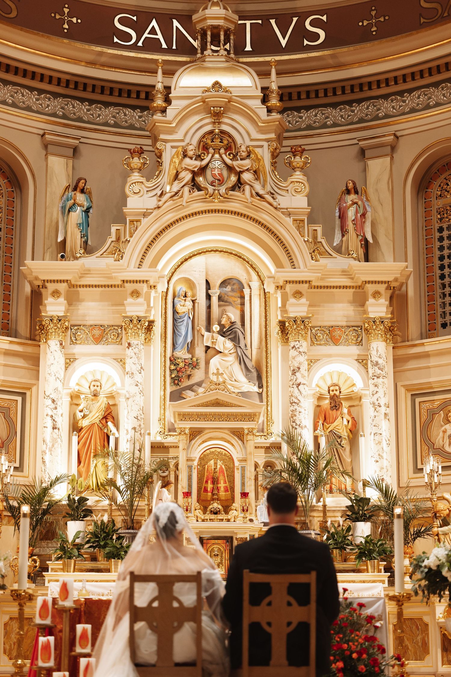 Bride and groom kneeling before ornate golden Catholic church altar during wedding ceremony.