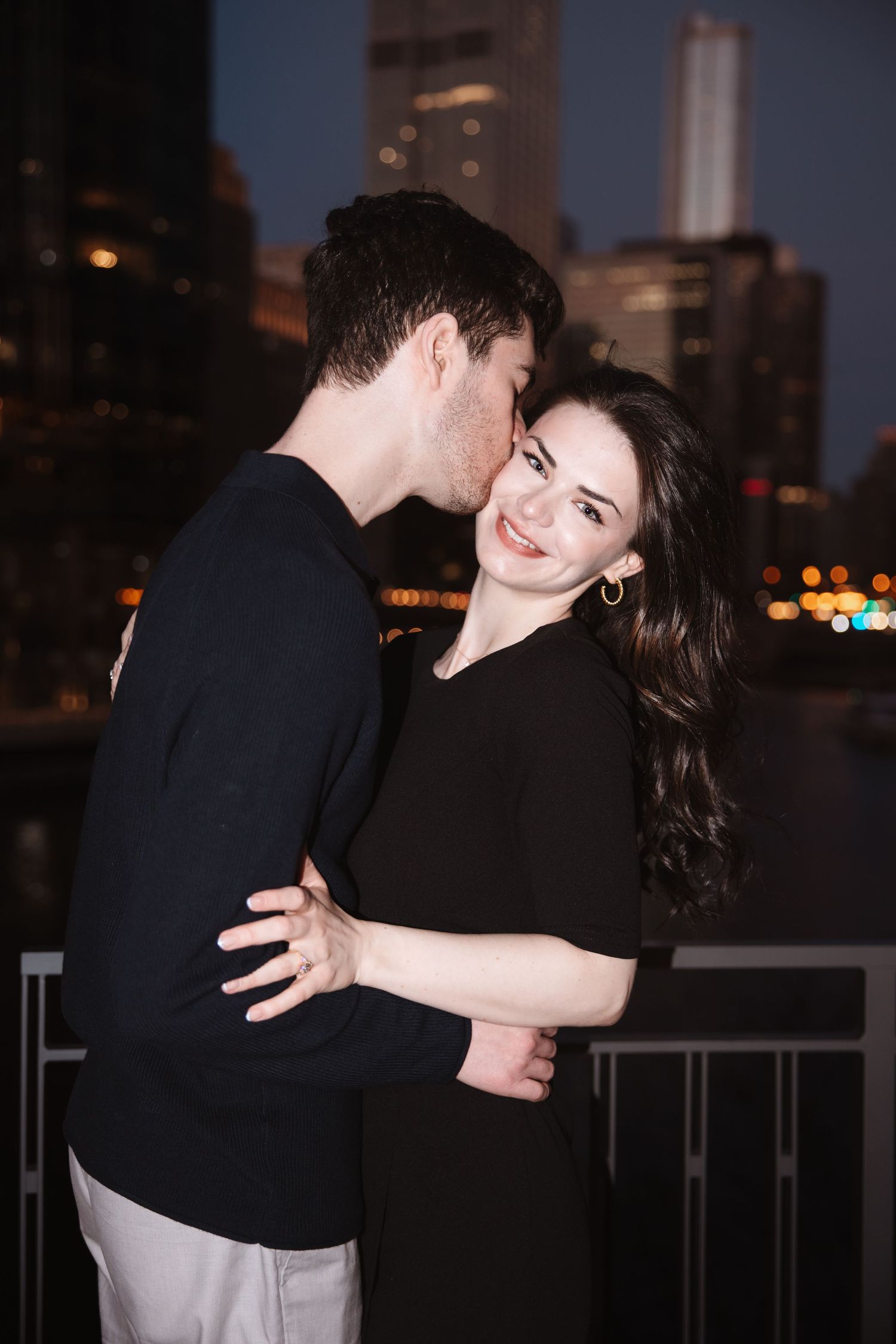 Happy woman smiles as her partner kisses her cheek during a romantic nighttime Chicago engagement session.