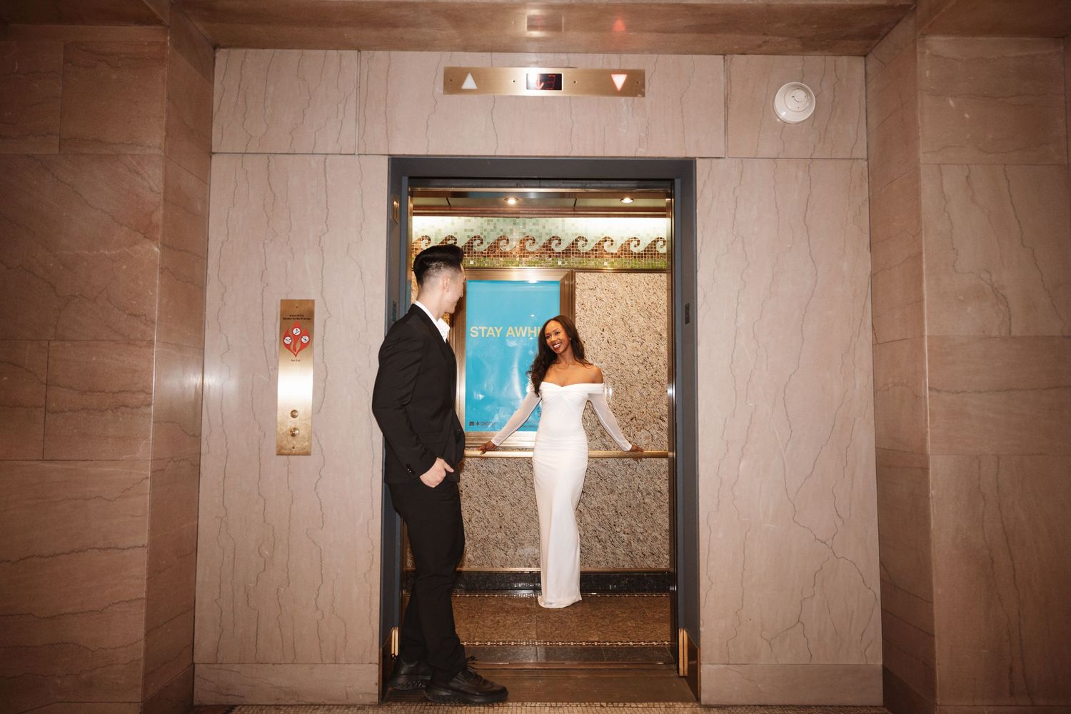 Interracial couple standing in an open elevator doorway, bride in white fitted gown and groom in black suit.Chicago Wedding Photographer