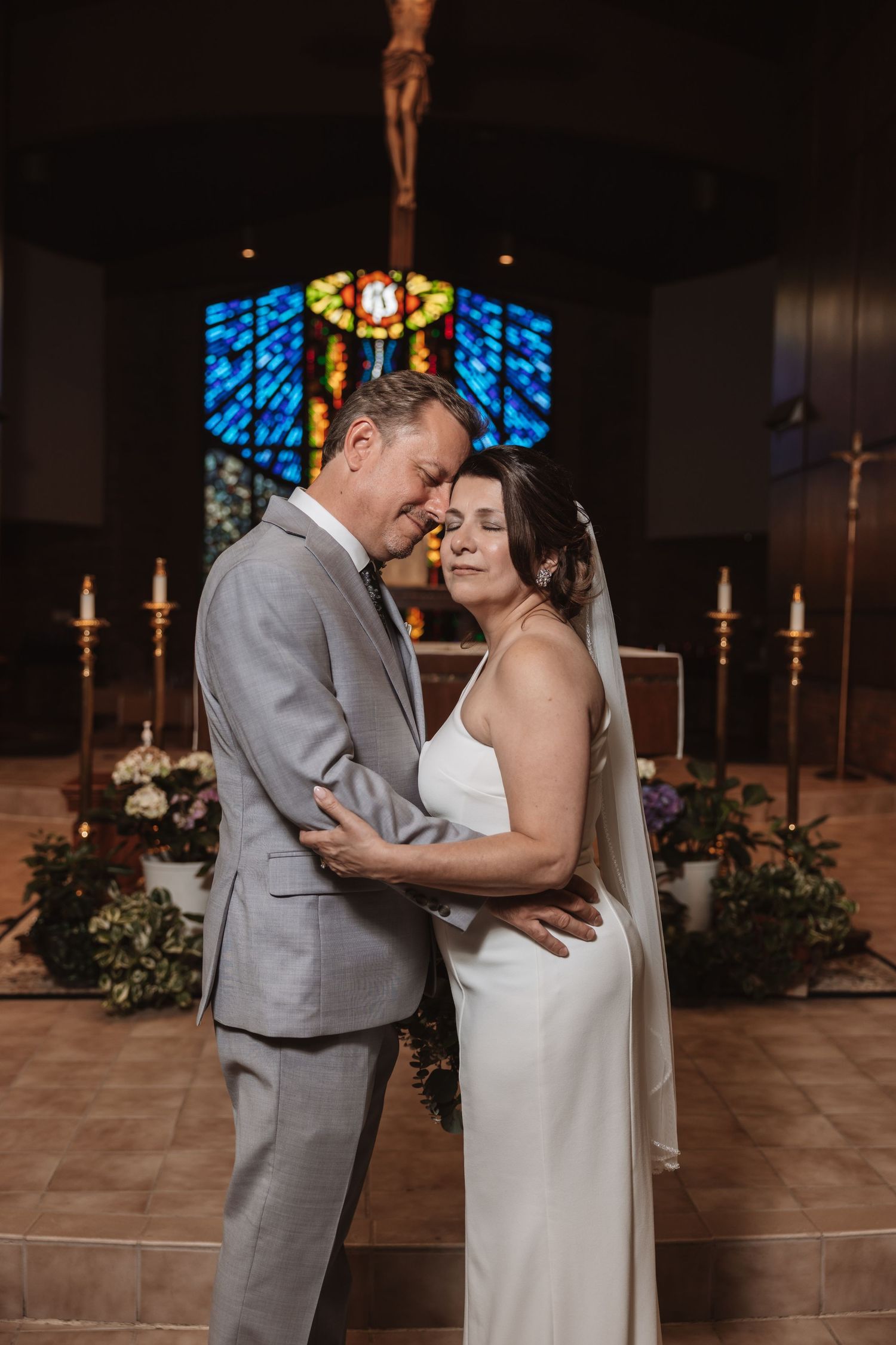 Bride and groom embrace tenderly at church altar with stained glass window and crucifix backdrop.