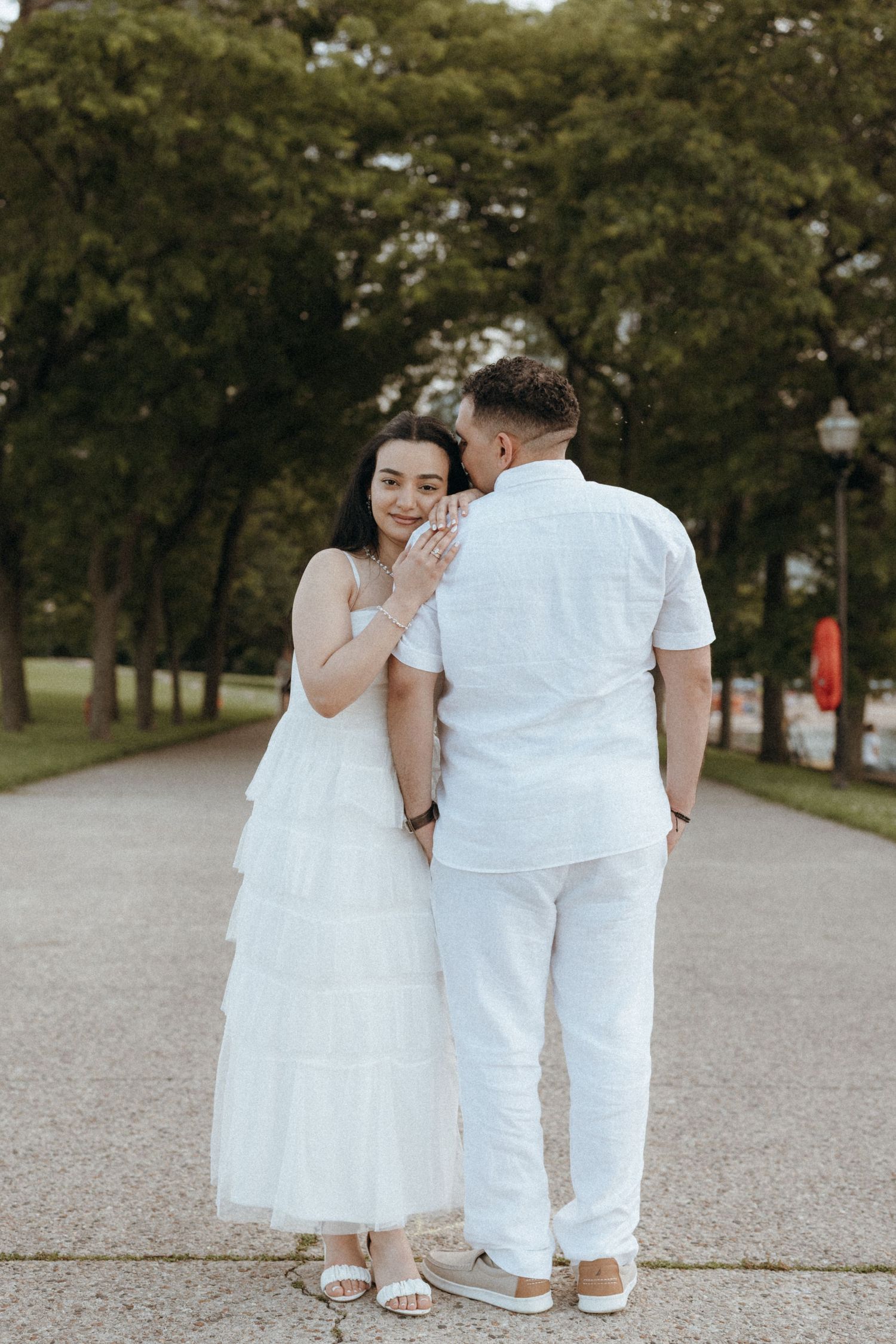 Couple in white outfits walking on a tree-lined path, man whispering to smiling woman.