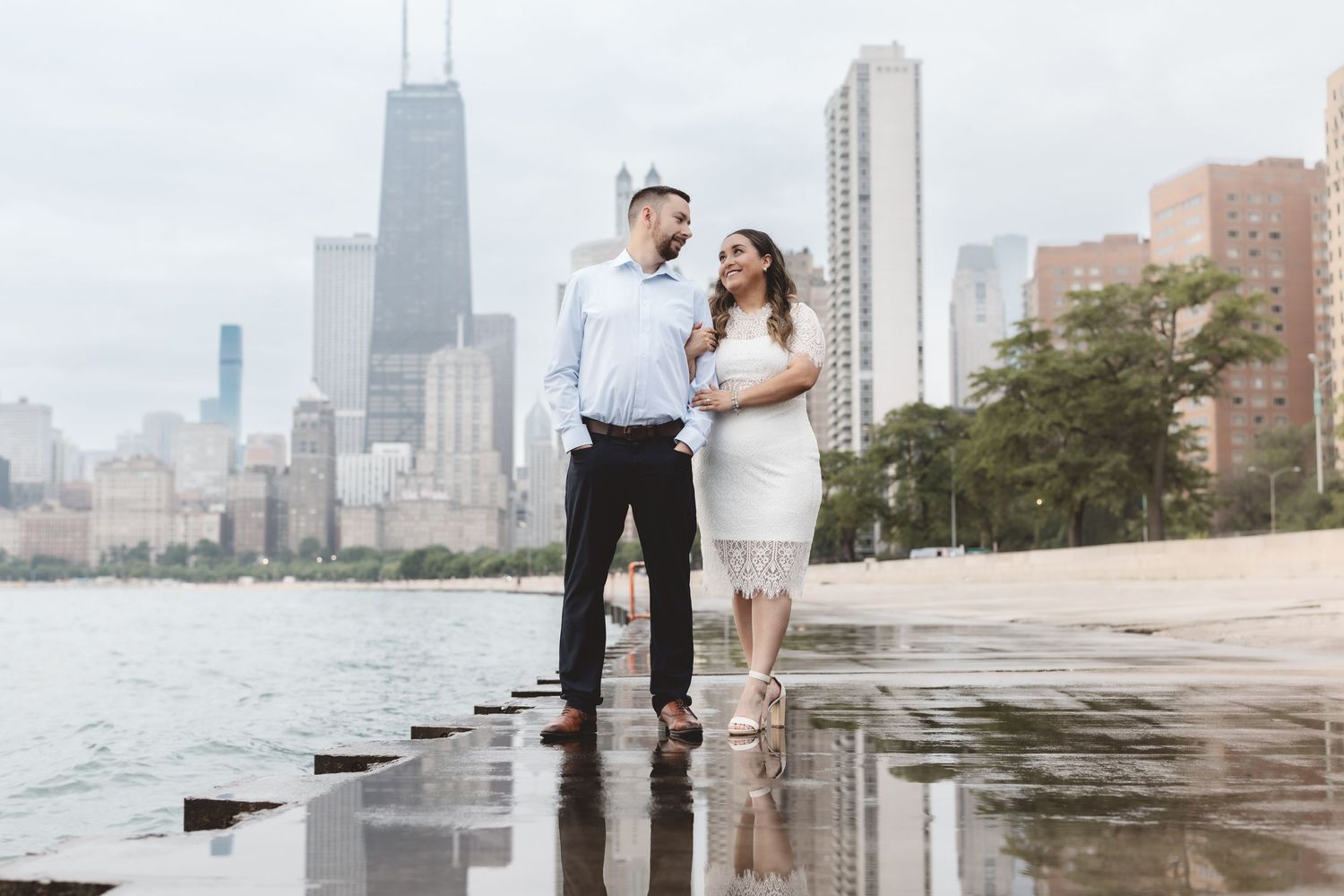 Couple walking along wet Chicago shoreline with skyline reflection and John Hancock Center behind them.
