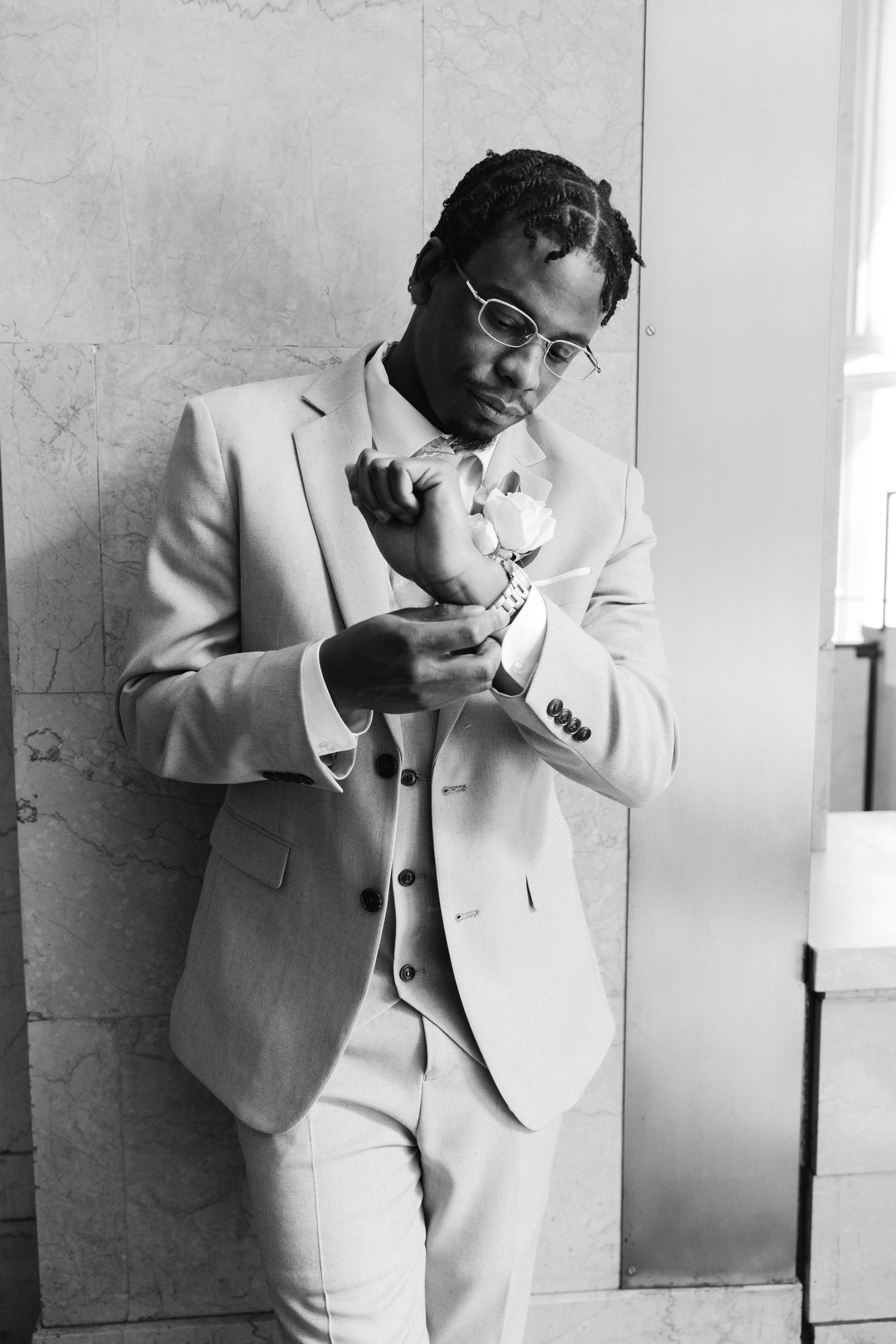Groom in light suit adjusts cufflinks while holding boutonniere in black and white wedding portrait.