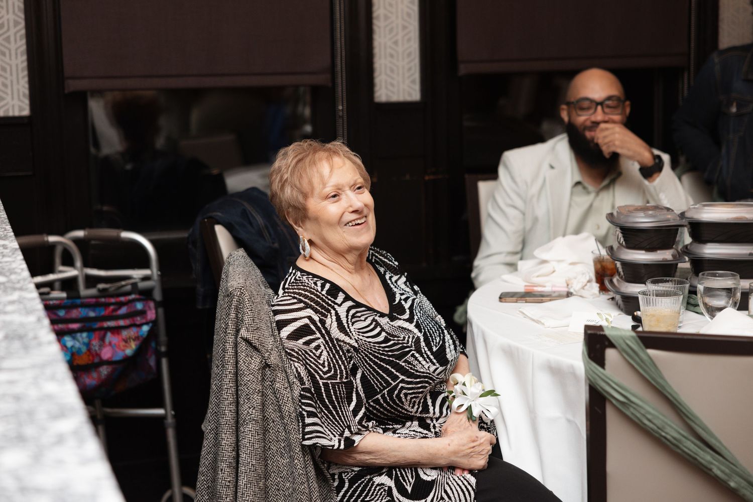 Elderly woman smiling joyfully at wedding reception table with guest in background.