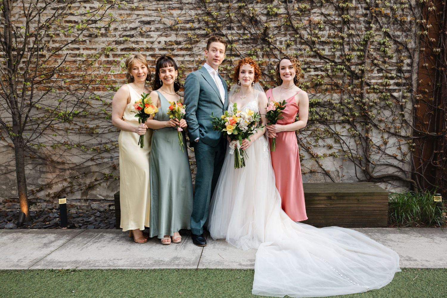Bride with long train and groom pose with three bridesmaids in pastel dresses against an ivy-covered brick wall.