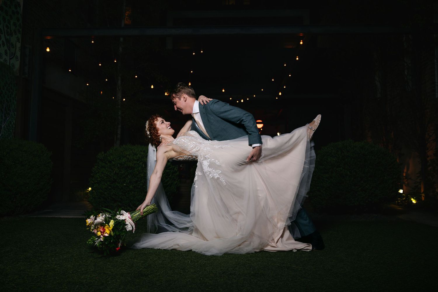 Bride and groom share a romantic dip kiss outdoors at night, surrounded by twinkling string lights.