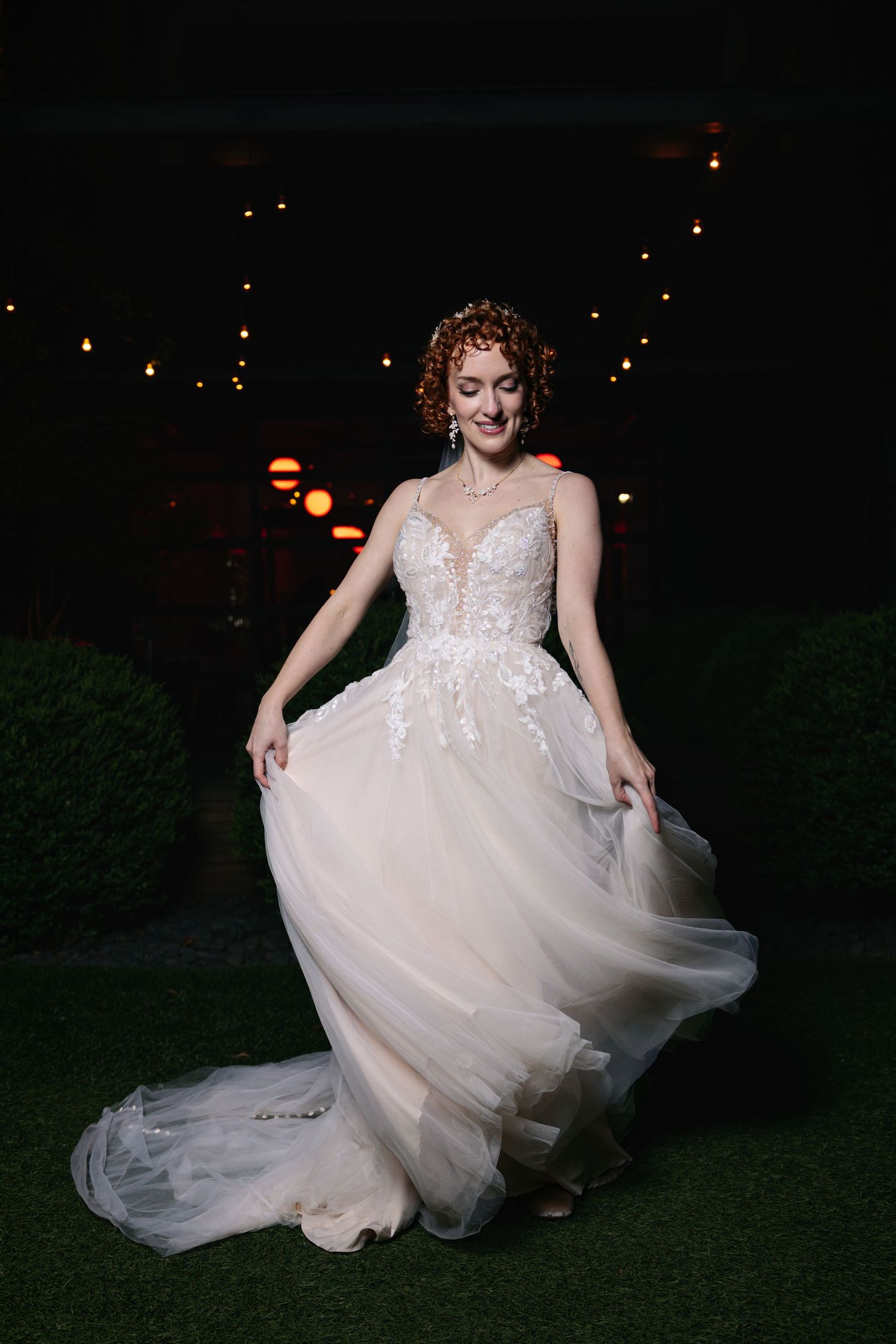 Bride in lace tulle ballgown twirling outdoors at night with string lights glowing in background.