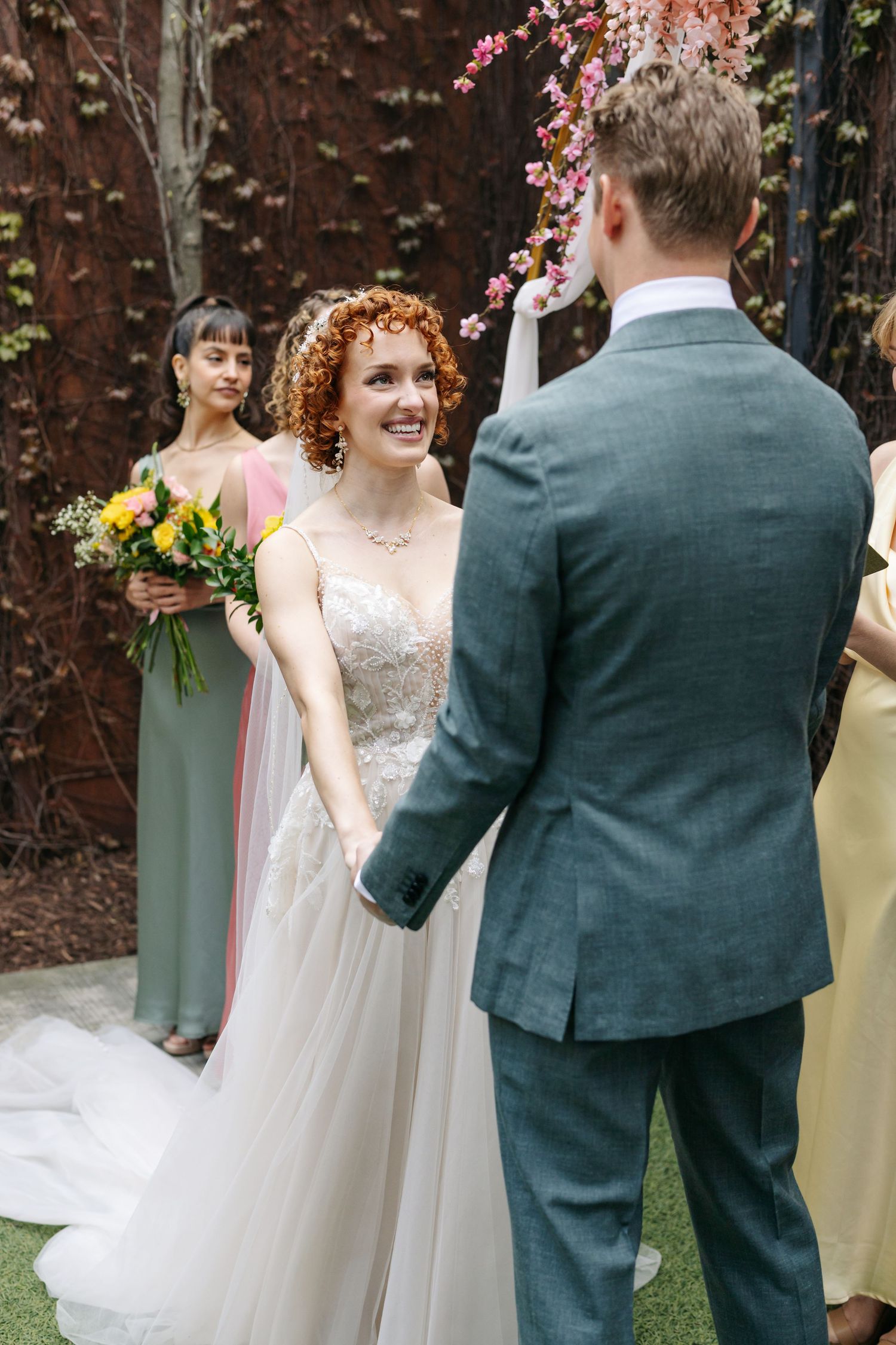 Smiling redhead bride holds hands with groom at outdoor wedding ceremony with bridesmaids behind.