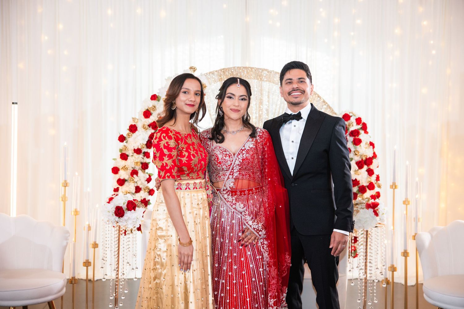 Indian bride in red lehenga stands between two guests at decorated wedding reception backdrop.