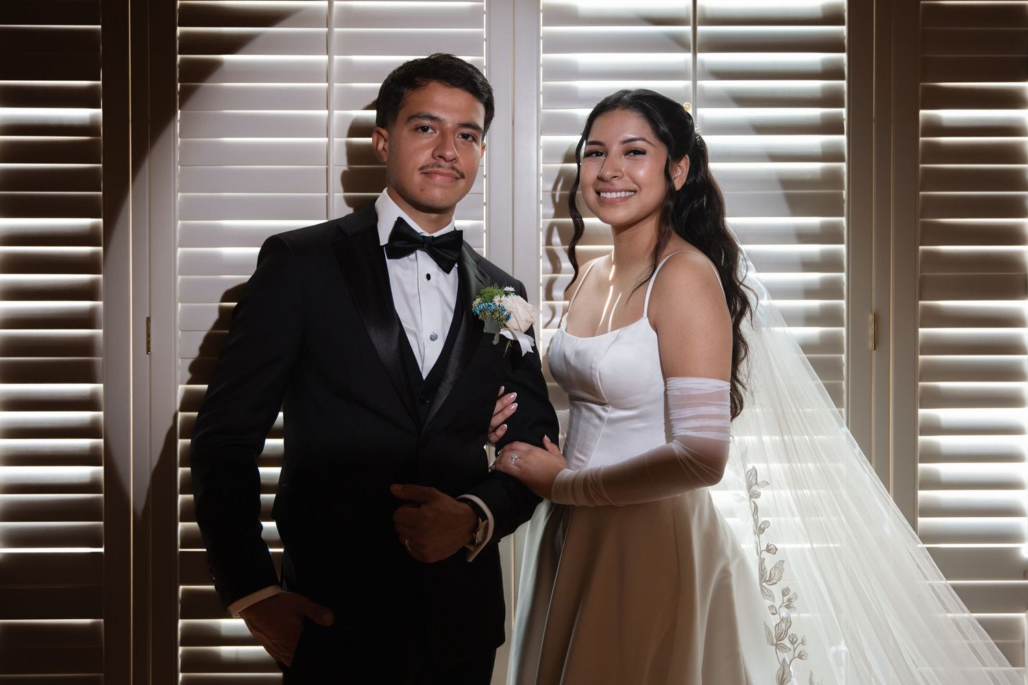 Smiling bride and groom posing together indoors, bride wearing veil and long white gloves on wedding day.