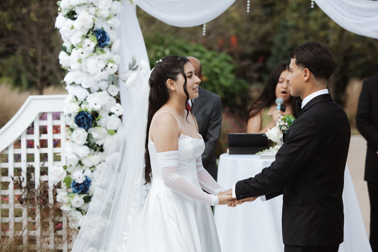Laughing bride and groom hold hands at outdoor wedding with white and blue floral arch.