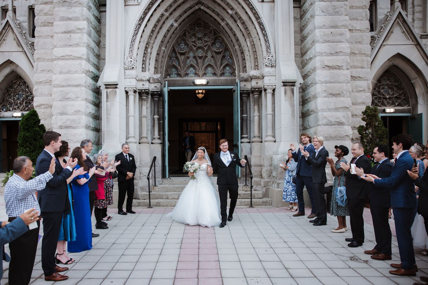 Happy bride and groom emerge from gothic church entrance as wedding guests cheer and celebrate outside.
