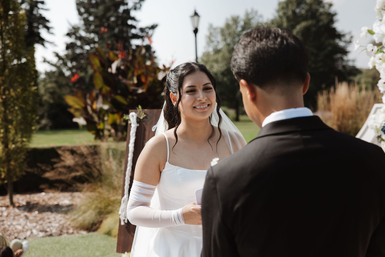 Smiling bride in white dress and gloves holds grooms hands during outdoor wedding ceremony.