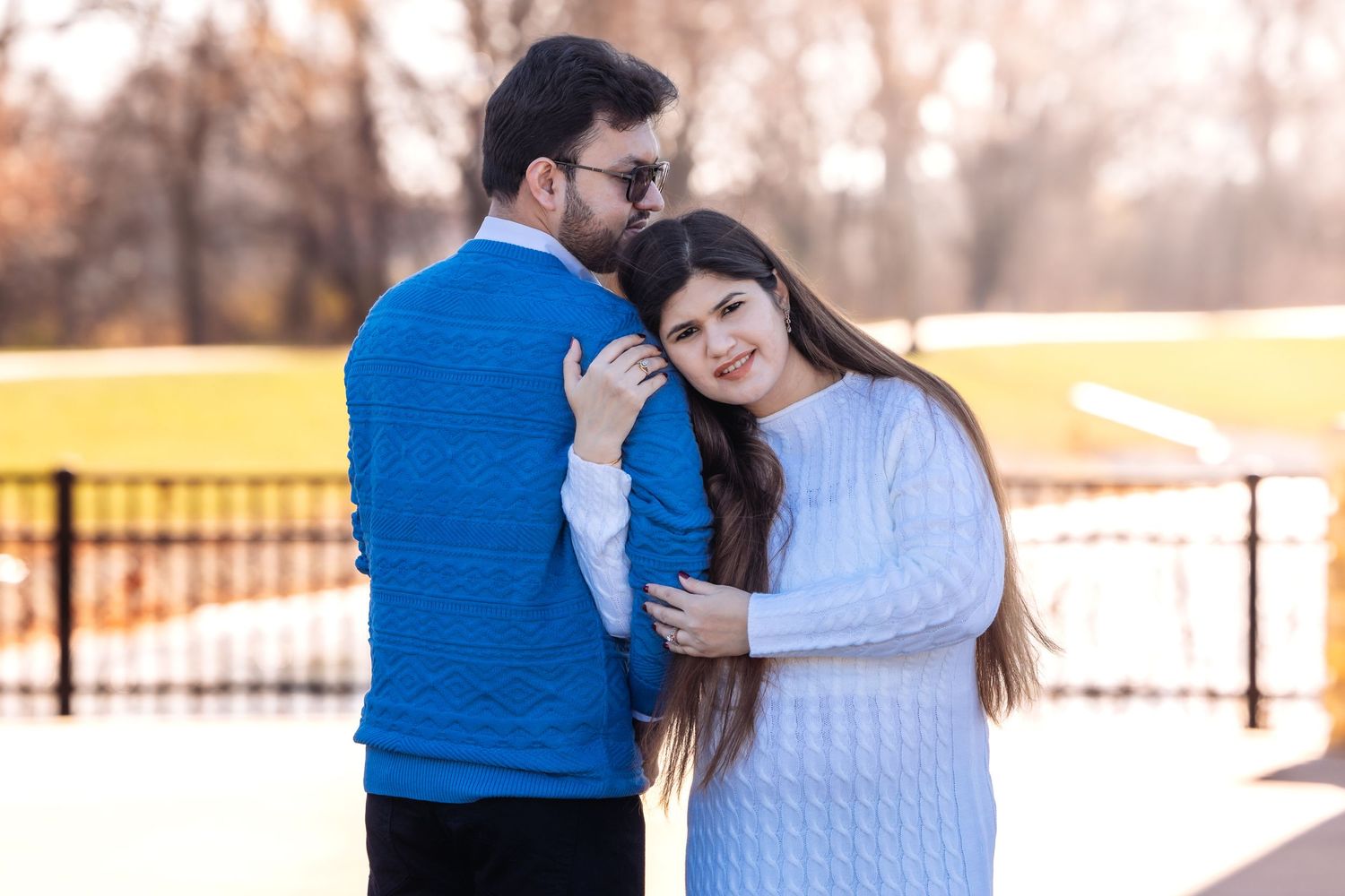 Couple embracing outdoors in park, man in blue sweater kissing woman's head, fall foliage background.