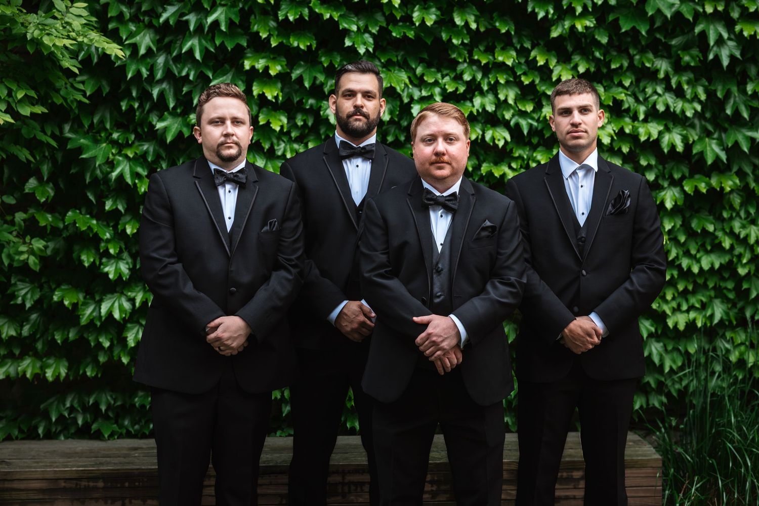 Four groomsmen in black tuxedos sitting and standing in front of a green ivy wall.