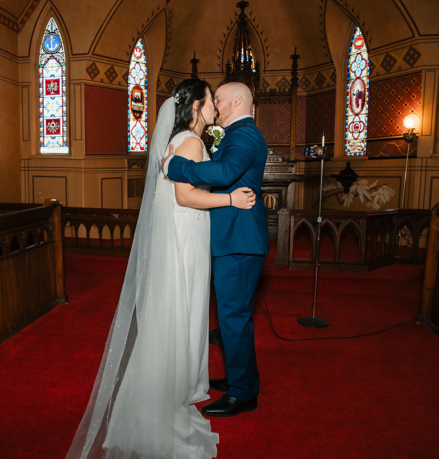 Bride and groom share first kiss at church altar with stained glass windows and red carpet aisle.