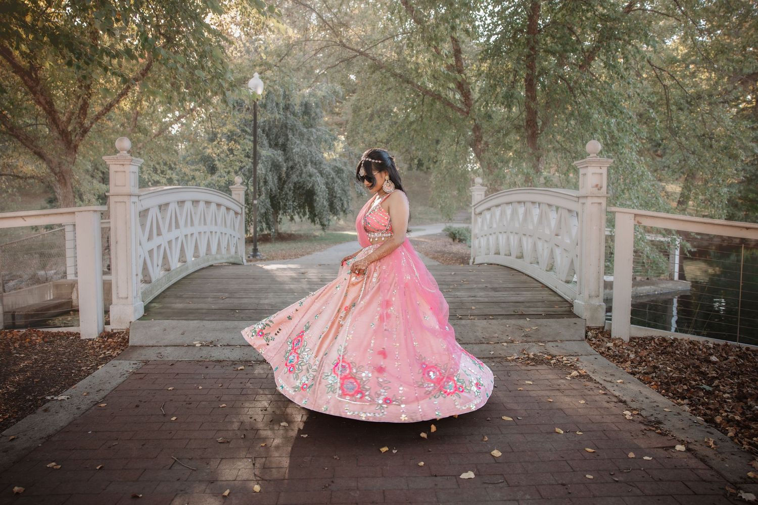 Indian bride in pink lehenga twirls gracefully on white bridge in lush garden setting.