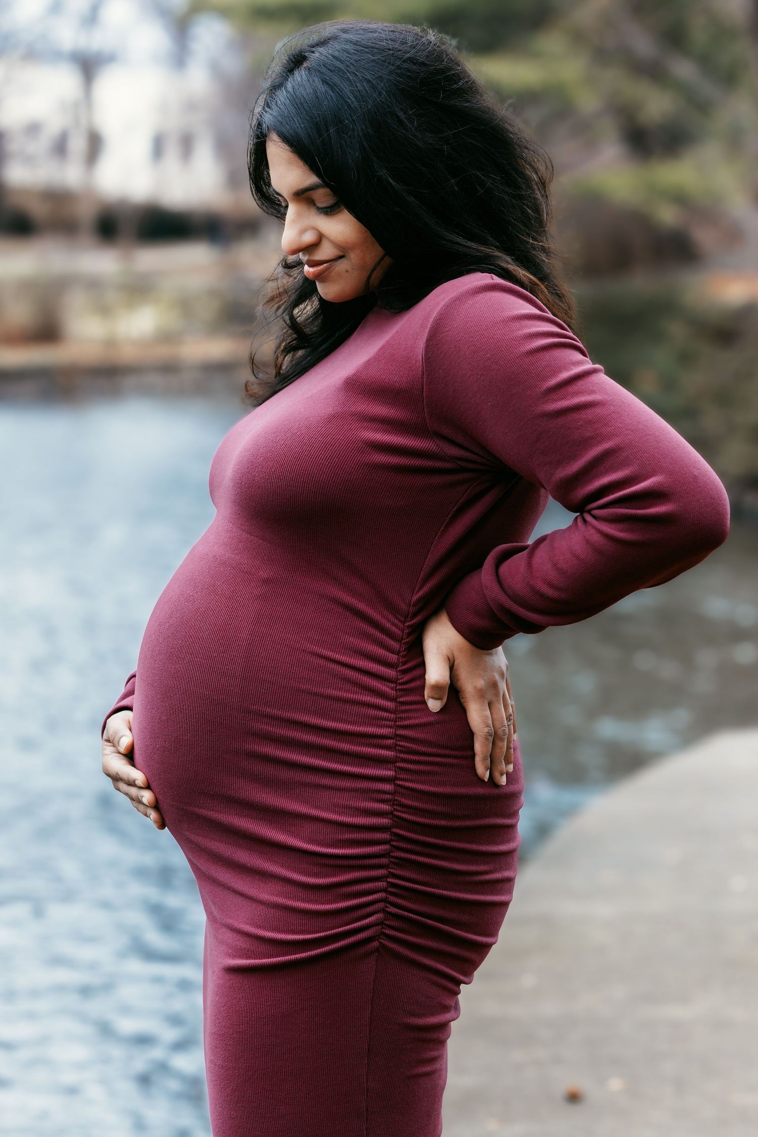 Pregnant woman in burgundy ruched dress posing sideways, cradling bump near a reflective waterway.