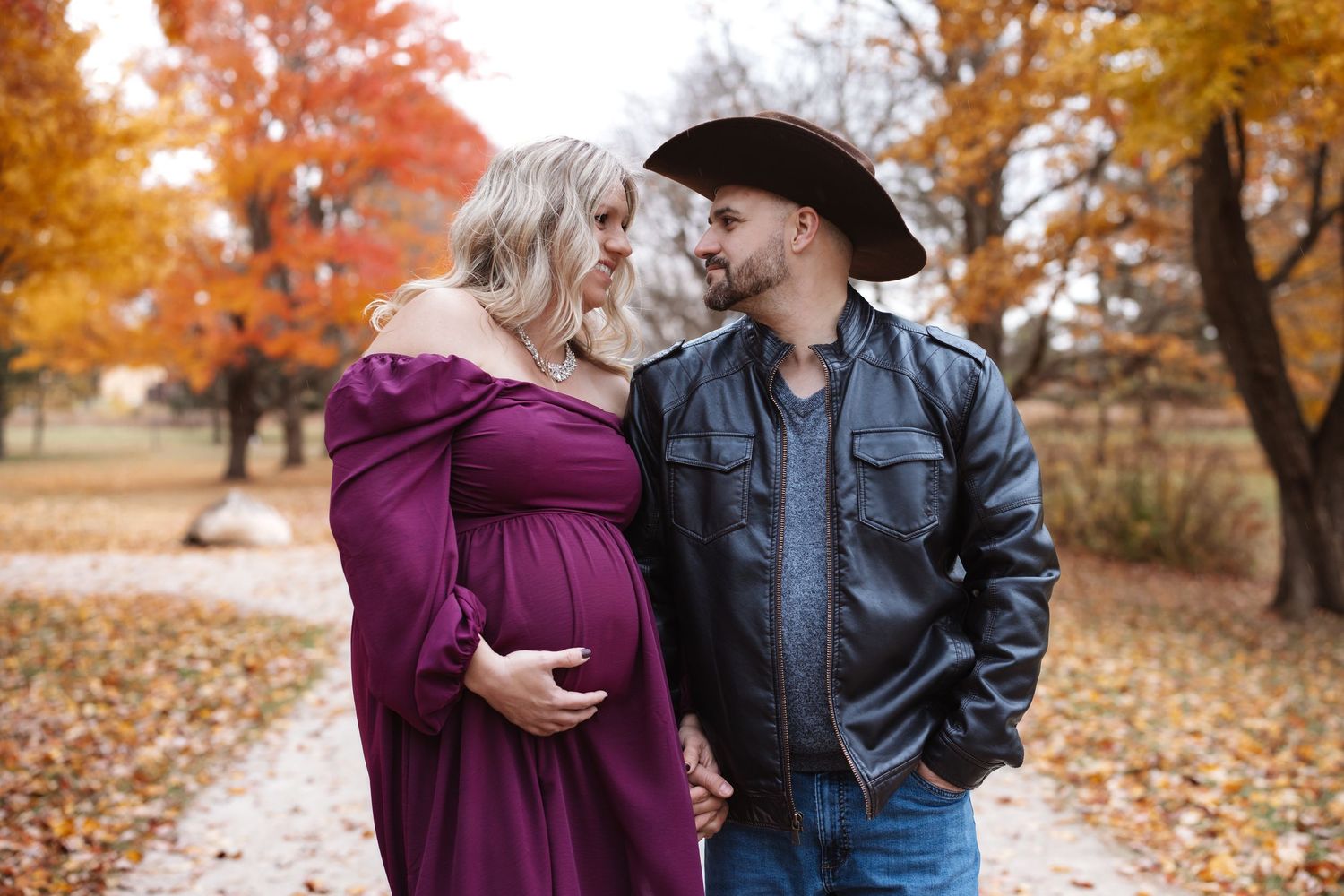 Pregnant couple sharing a tender moment on a scenic path surrounded by vibrant fall foliage.