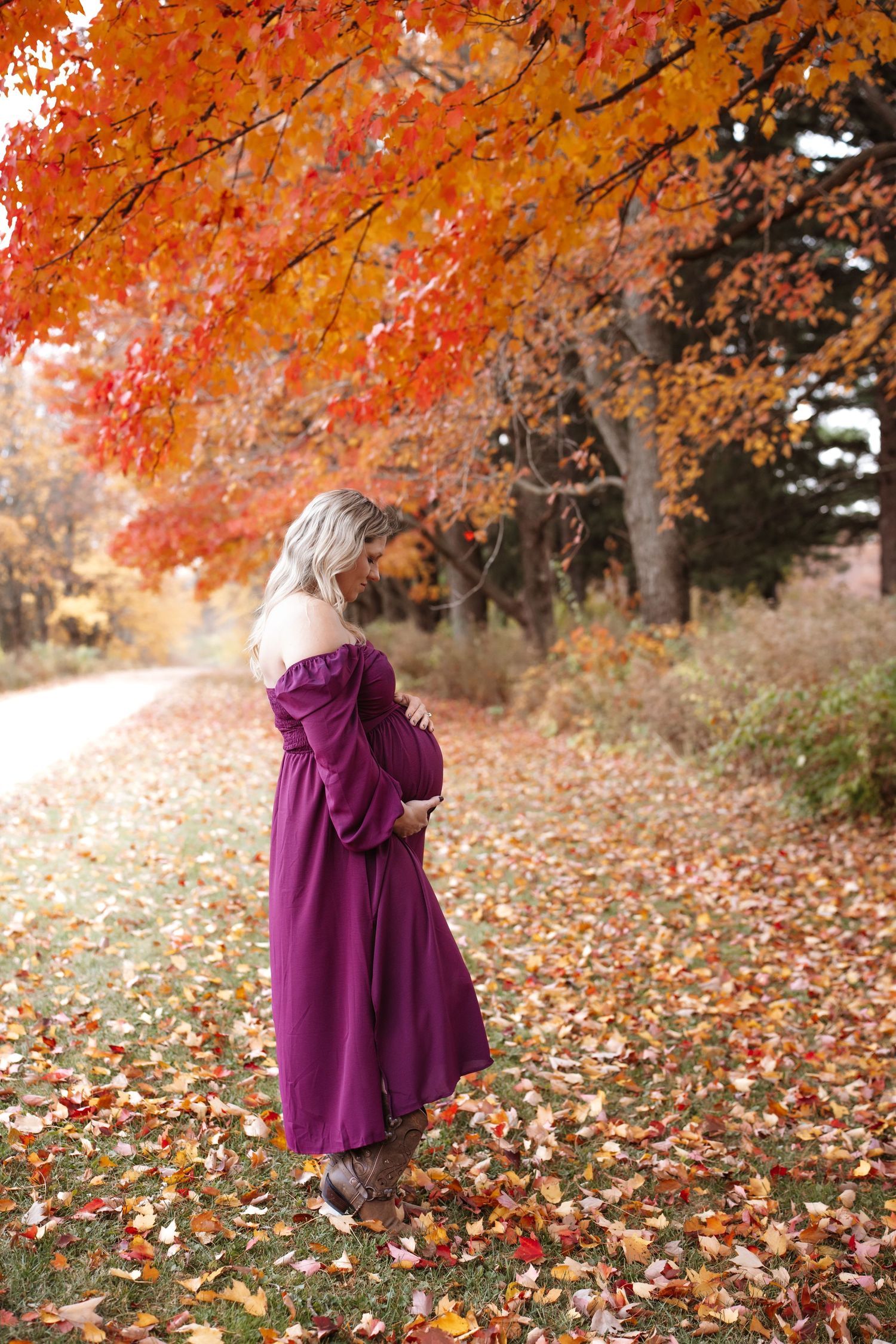 Pregnant woman in purple off-shoulder dress standing on leaf-covered ground beneath autumn tree canopy.