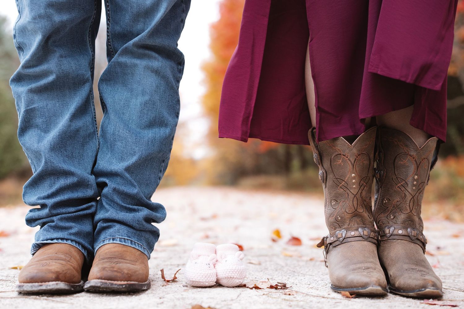 Close-up of couple in cowboy boots with tiny pink baby shoes between them on autumn path.