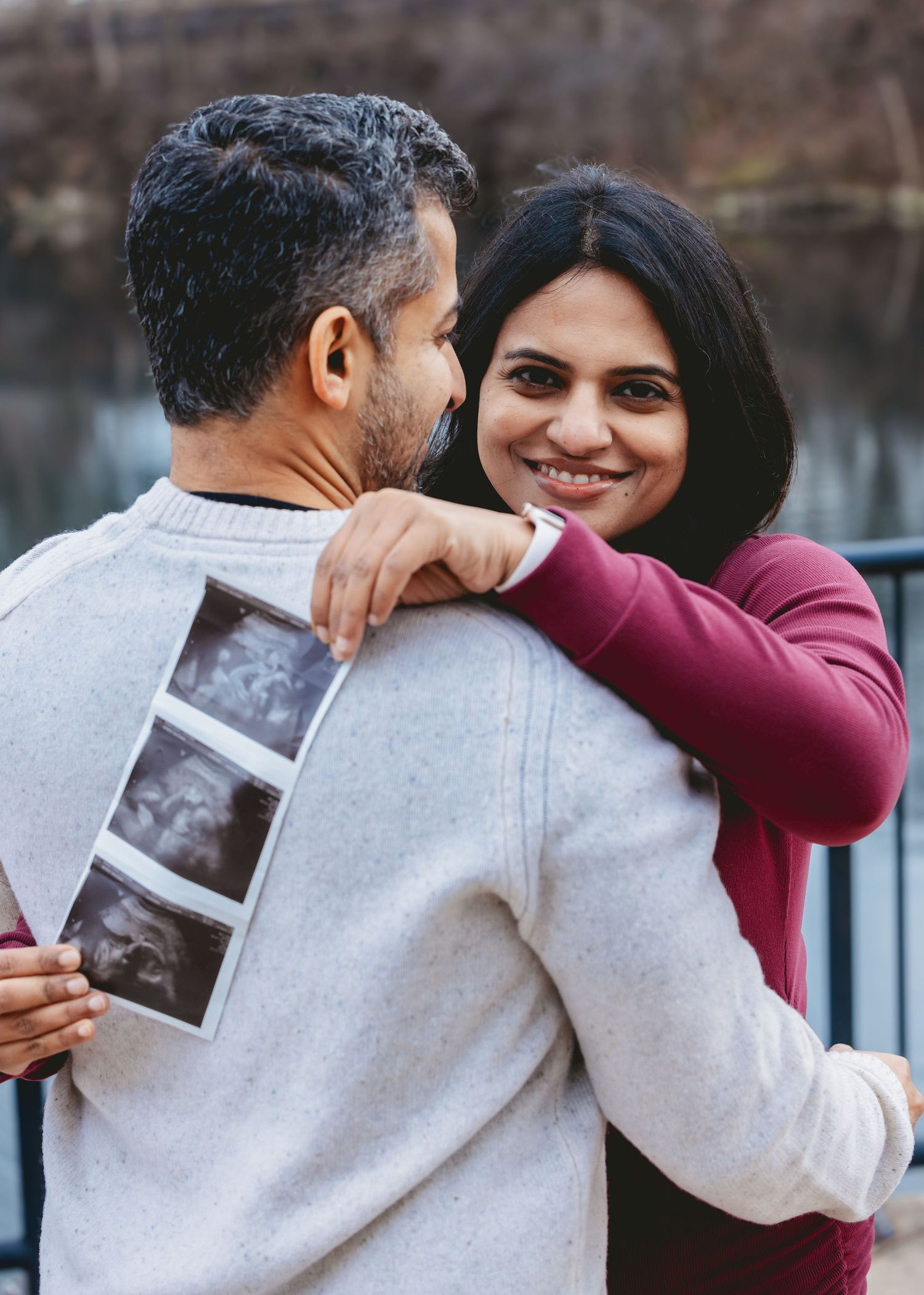 Couple embracing outdoors, woman smiling as man holds ultrasound photos behind his back near a lake.
