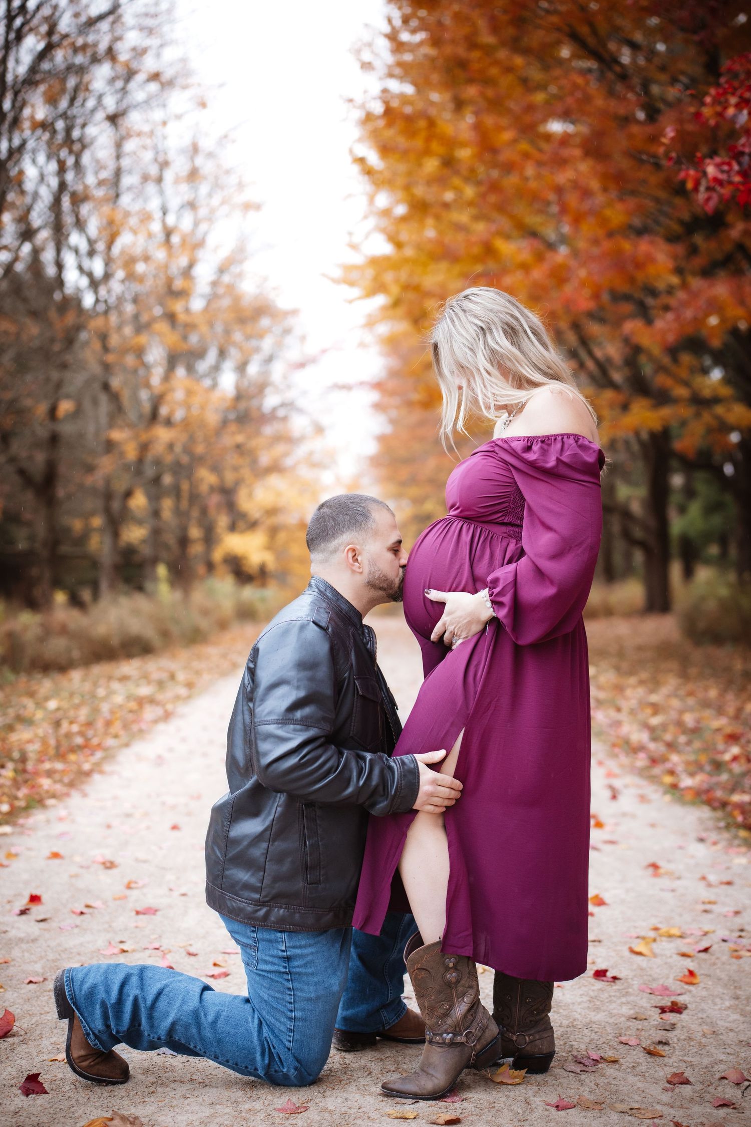 Man kneeling to kiss pregnant woman's belly on a leaf-covered path in autumn forest.