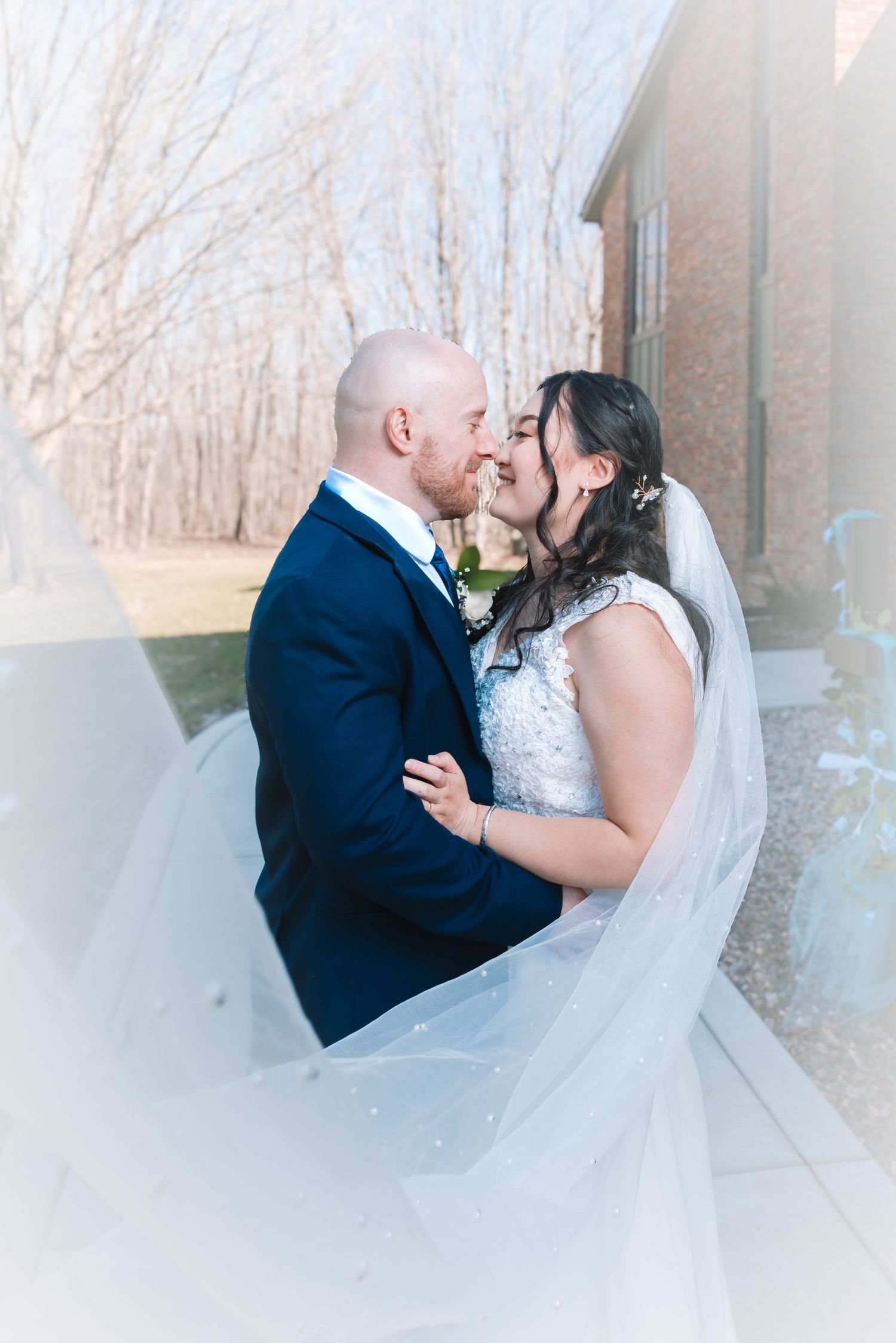 Bride and groom nearly kissing as flowing veil wraps around them near brick building.