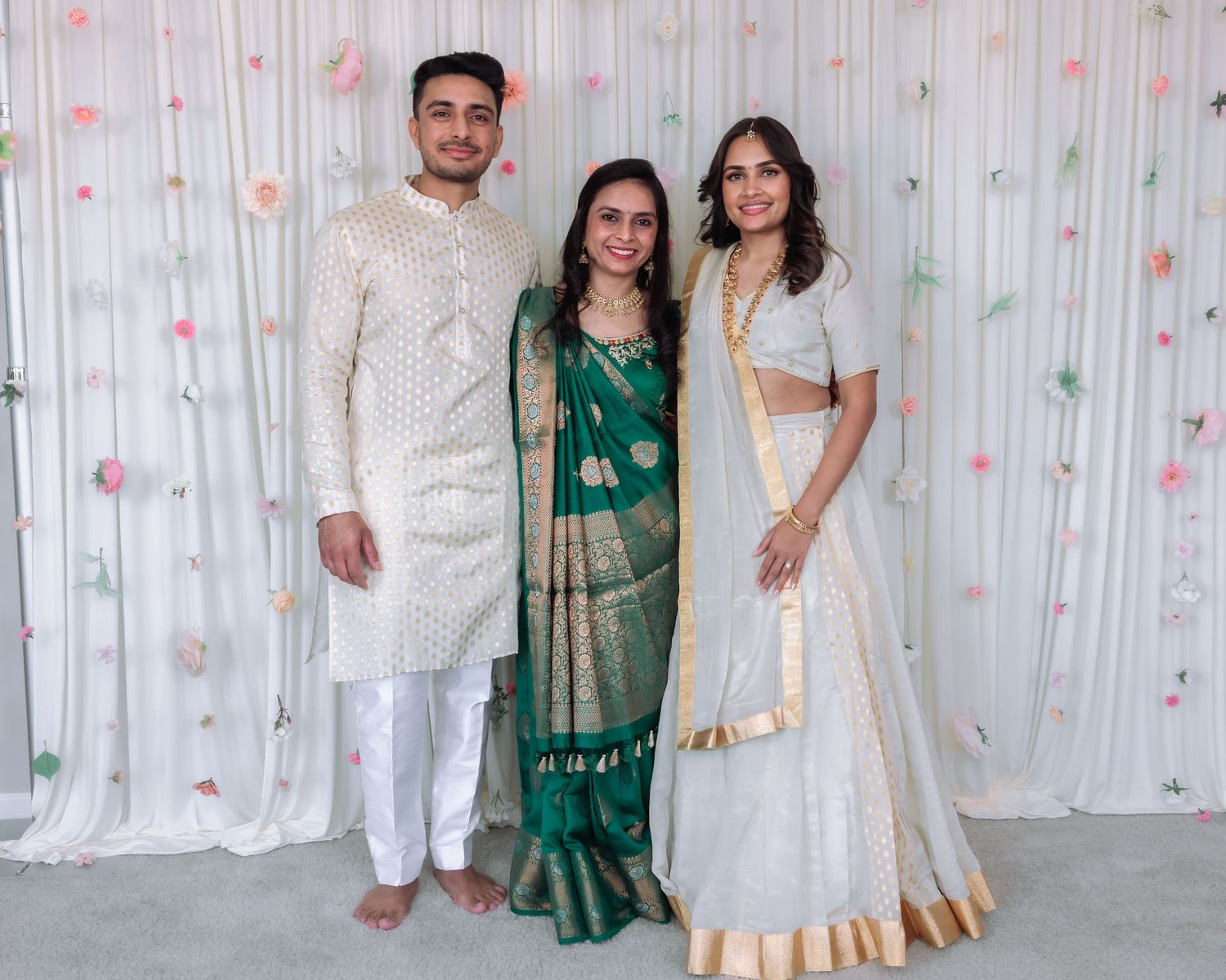 Three young adults in traditional Indian outfits, two in white and one in green saree, posing at a celebration.