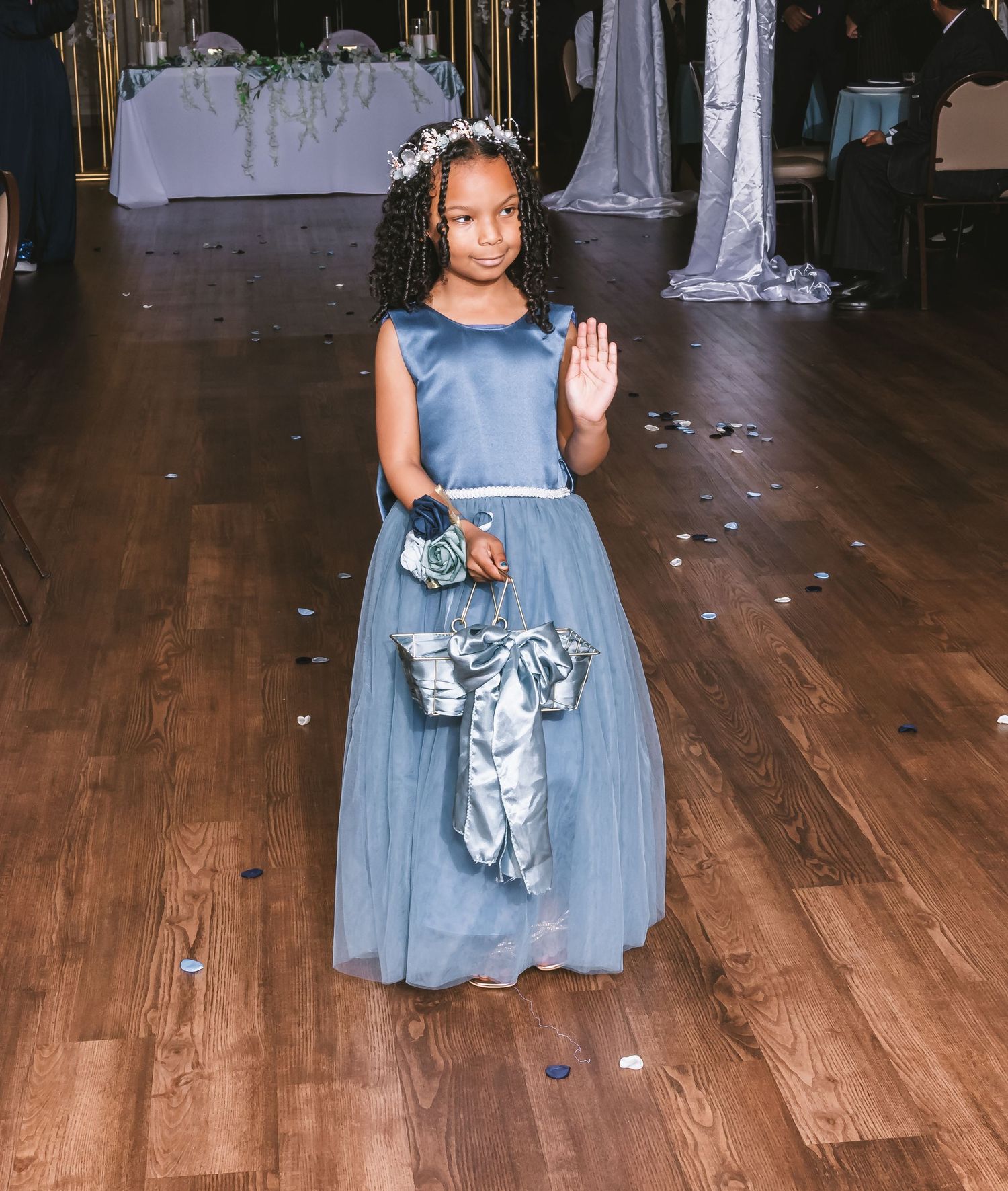 A young flower girl in a dusty blue dress waves while walking down the aisle at a wedding.