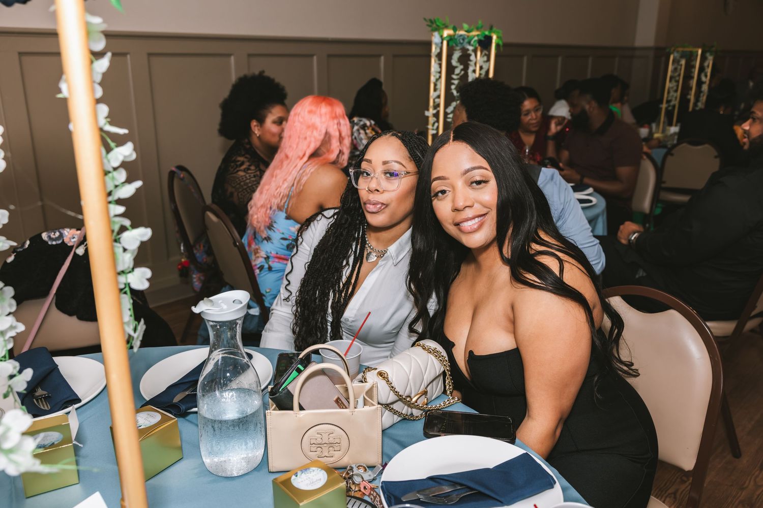 Two women smiling together at a wedding reception table decorated with white flowers and blue linens.