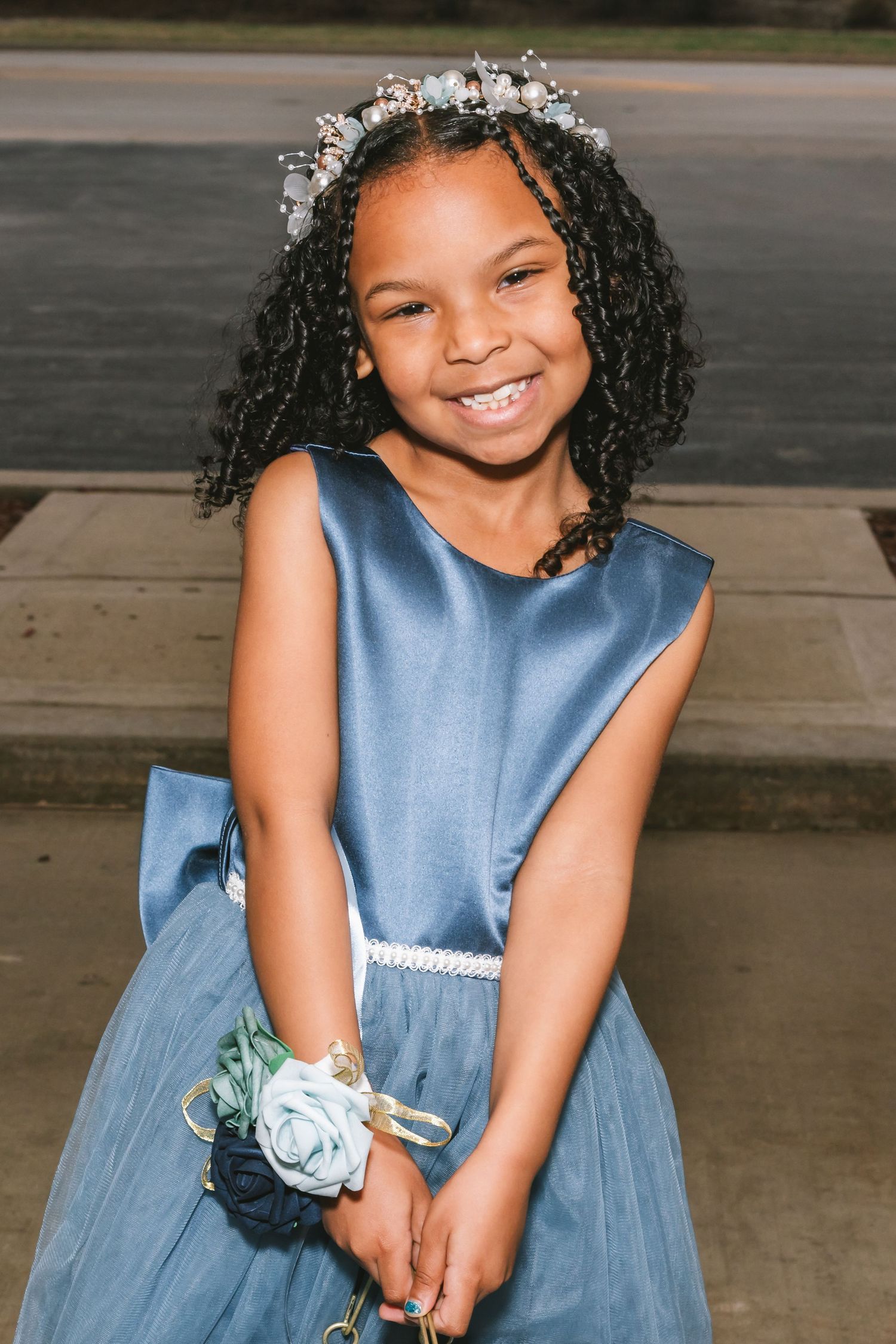 Adorable young flower girl in blue satin dress wearing floral crown smiling brightly at wedding celebration.