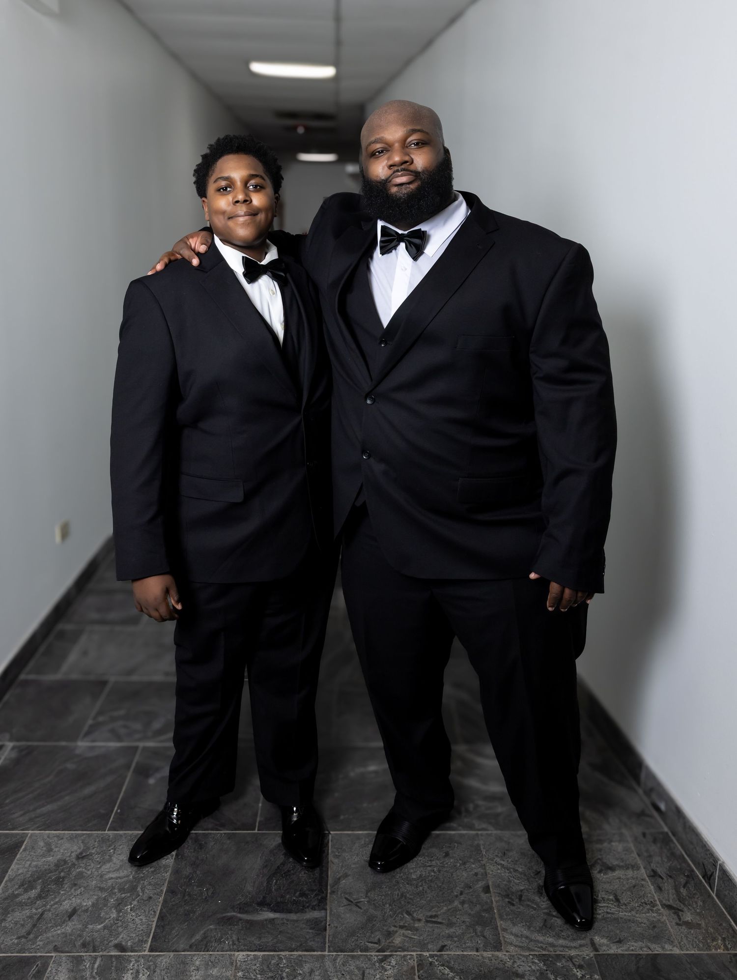 Two males in matching black tuxedos with bow ties posing together in a hallway.