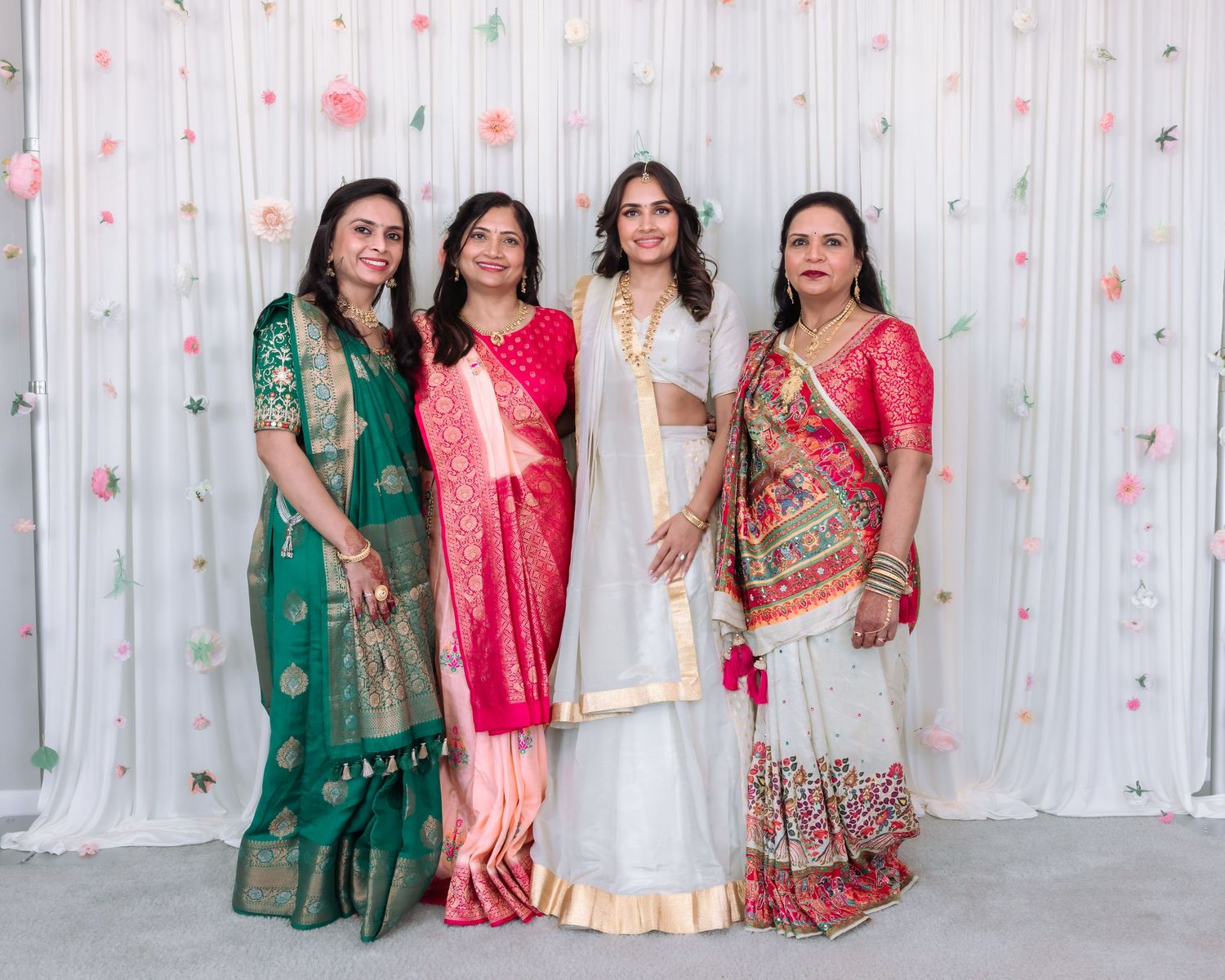 Four women wearing vibrant Indian traditional sarees and lehengas smile together at a floral decorated wedding backdrop.