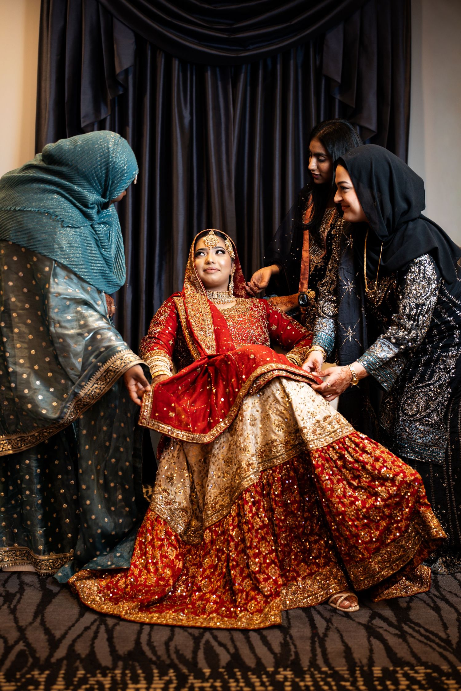 Bride in red gold lehenga laughing joyfully while three women in hijabs help arrange her outfit.