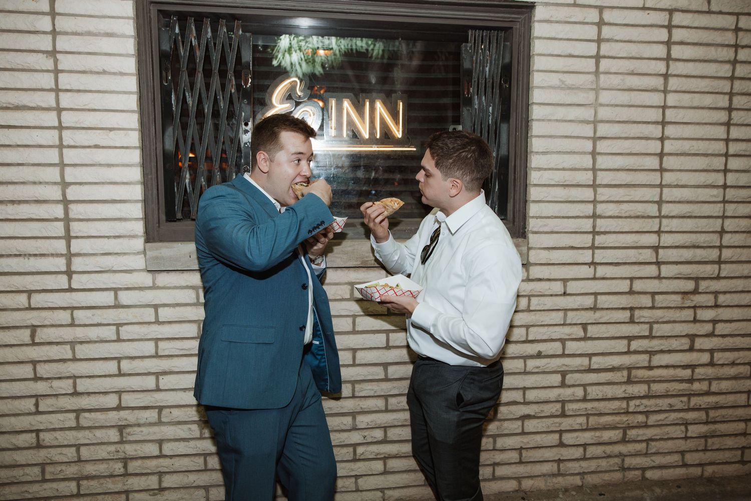 Two men in suits sharing food and laughing outside a lit inn sign at night.