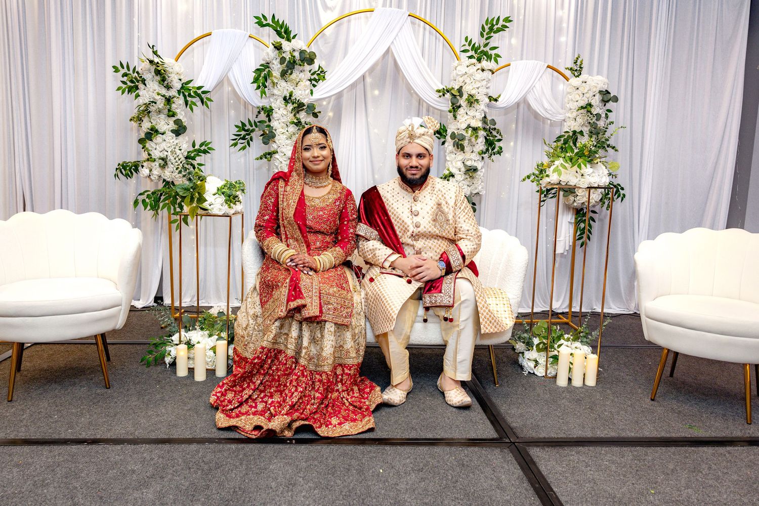 South Asian bride and groom in traditional red and gold wedding attire seated before elegant floral arch backdrop.