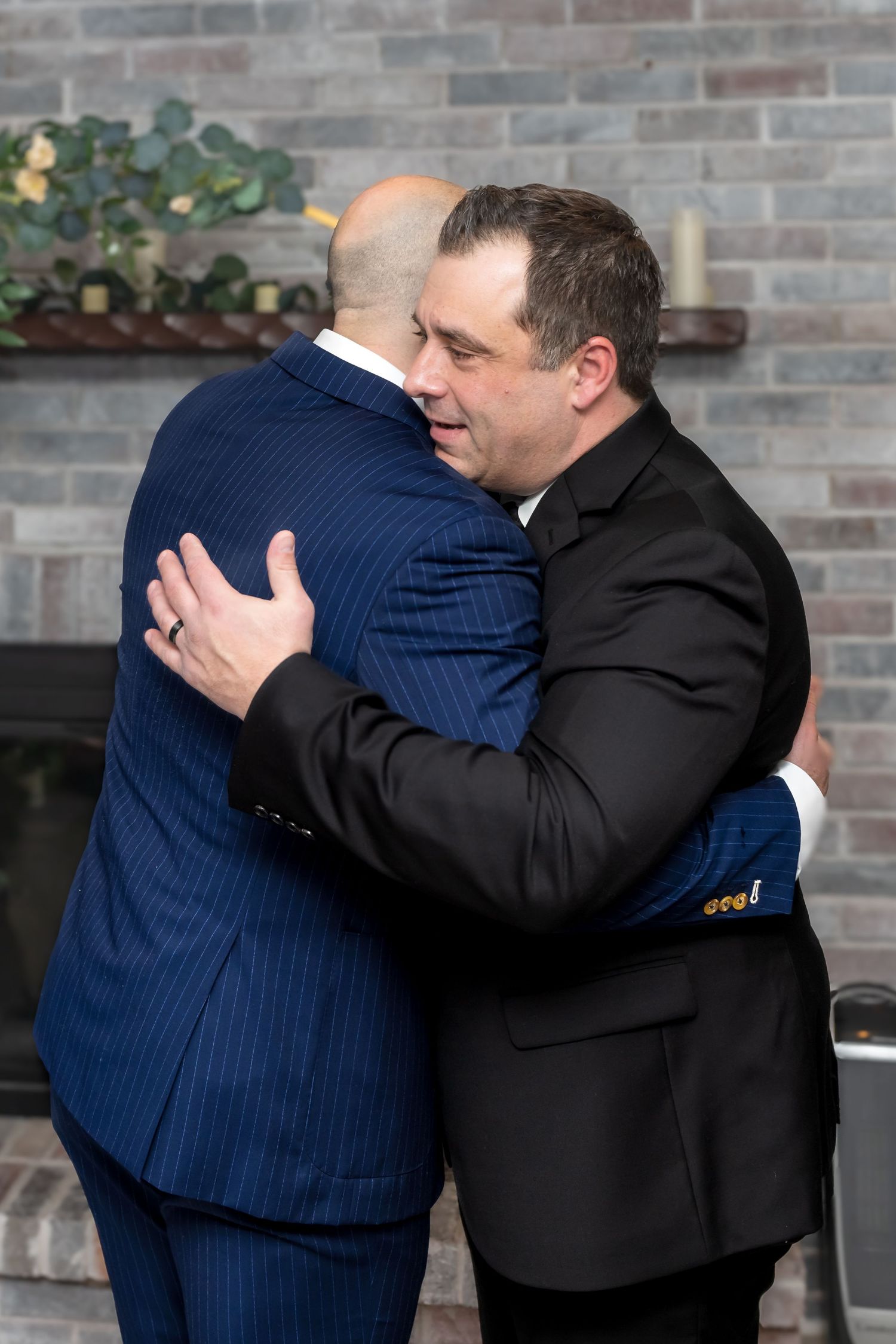 Two men in suits sharing an emotional hug in front of a brick fireplace.
