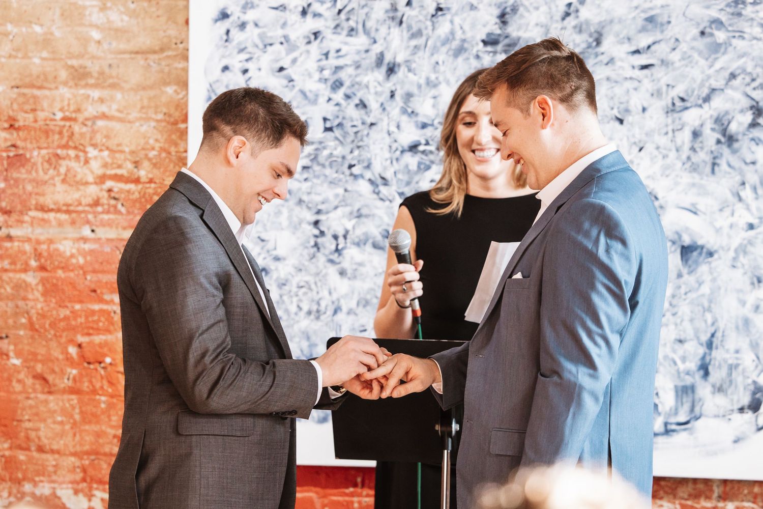 Two grooms exchange wedding rings while smiling with their female officiant in an art gallery ceremony.