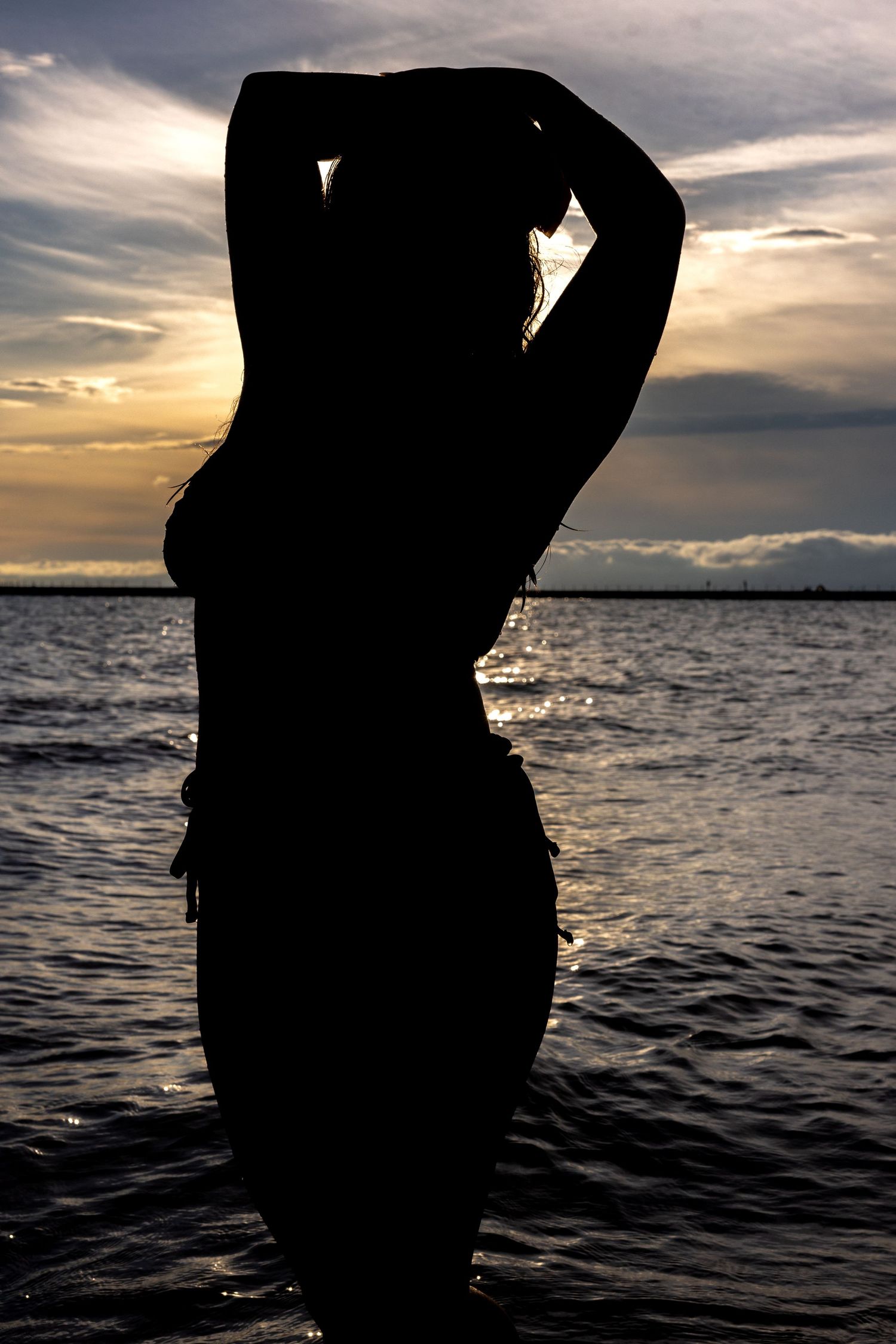 Silhouette of a woman with arms raised standing in ocean water at sunset.