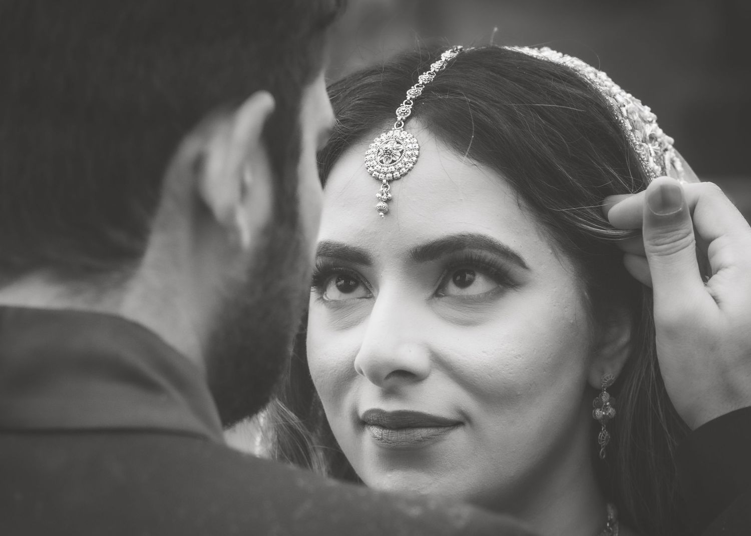 Black and white close-up of South Asian bride smiling as groom adjusts her maang tikka bridal headpiece.