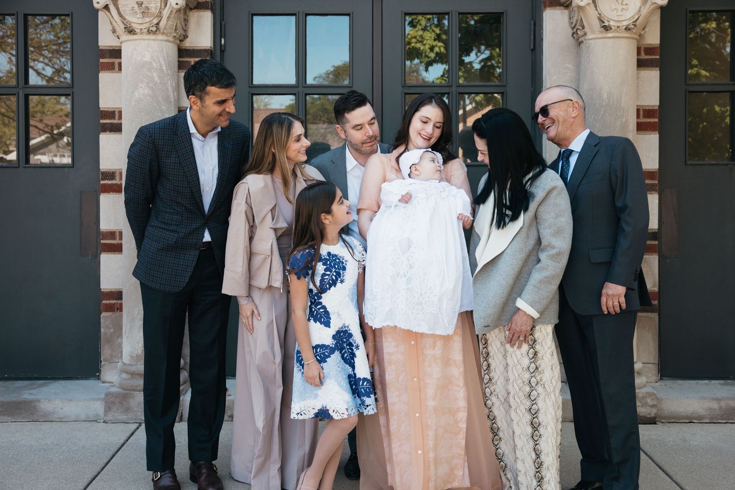 Extended family posing together with baby in white christening outfit outside church building.