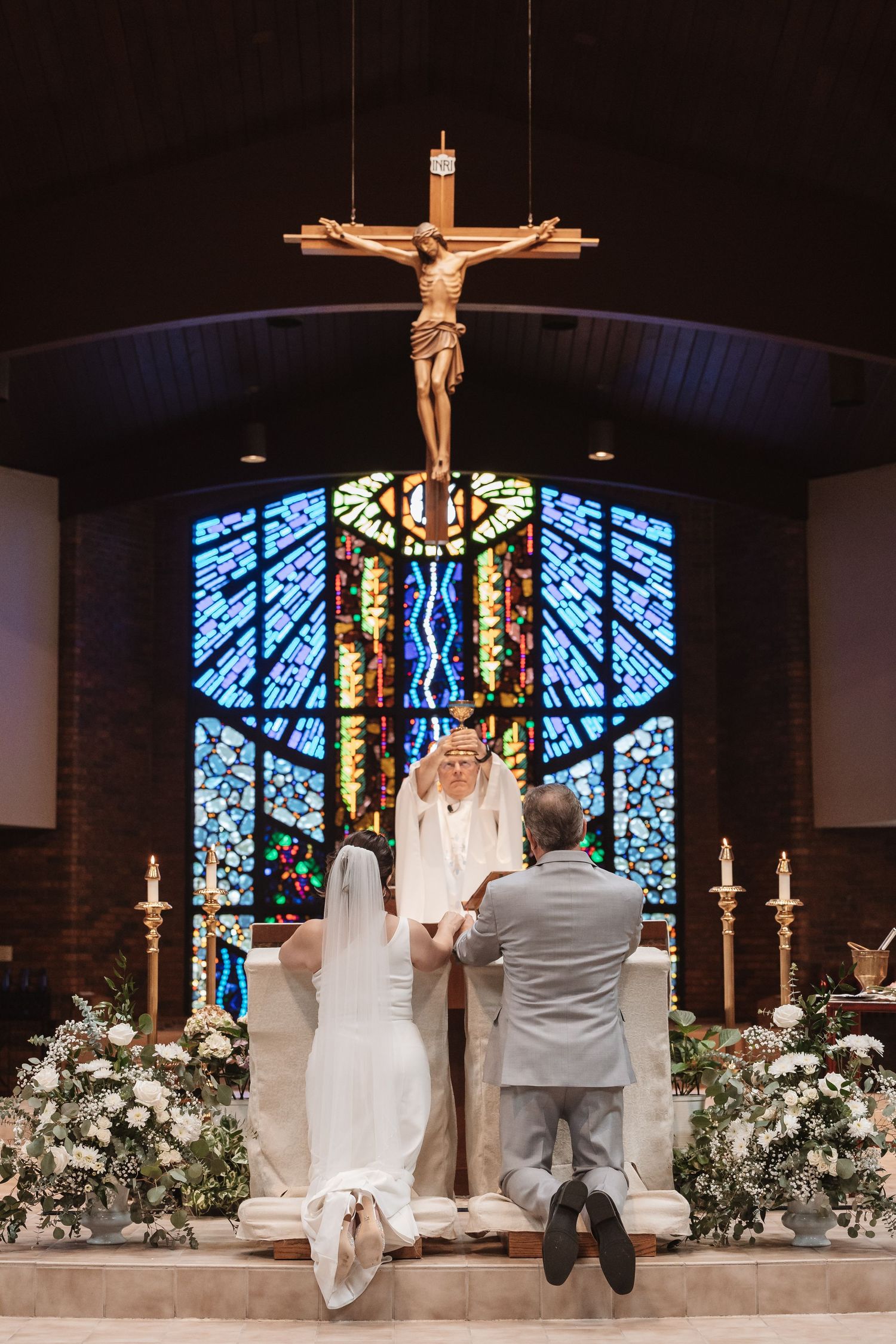 Bride and groom kneel at church altar beneath crucifix during Catholic wedding ceremony with stained glass windows.