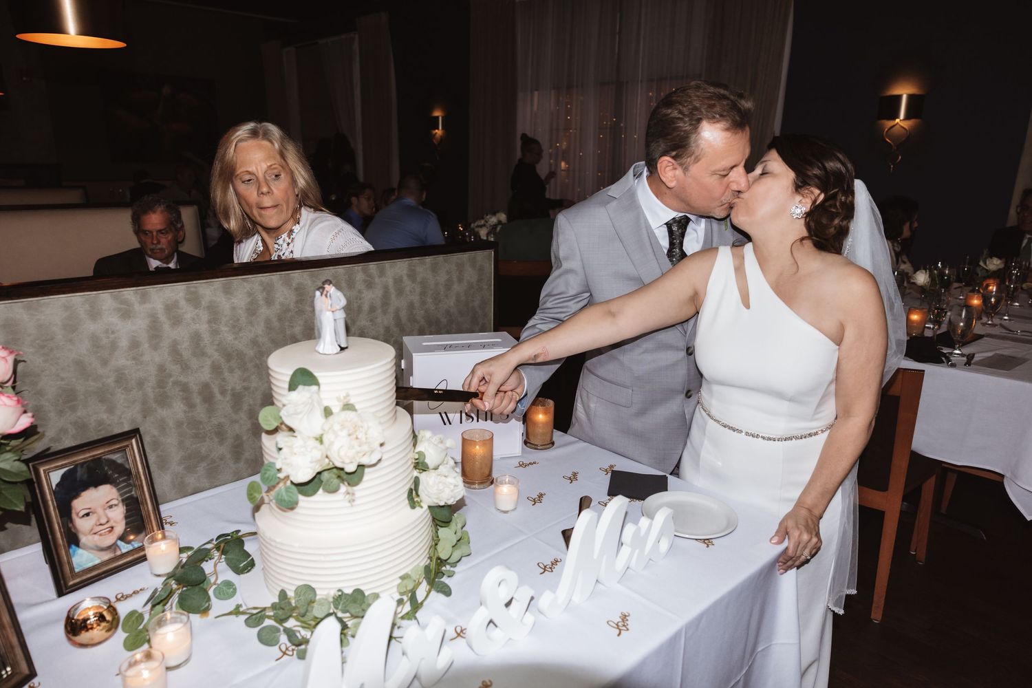 Bride and groom kiss while cutting white wedding cake at elegant reception dinner table with candles.