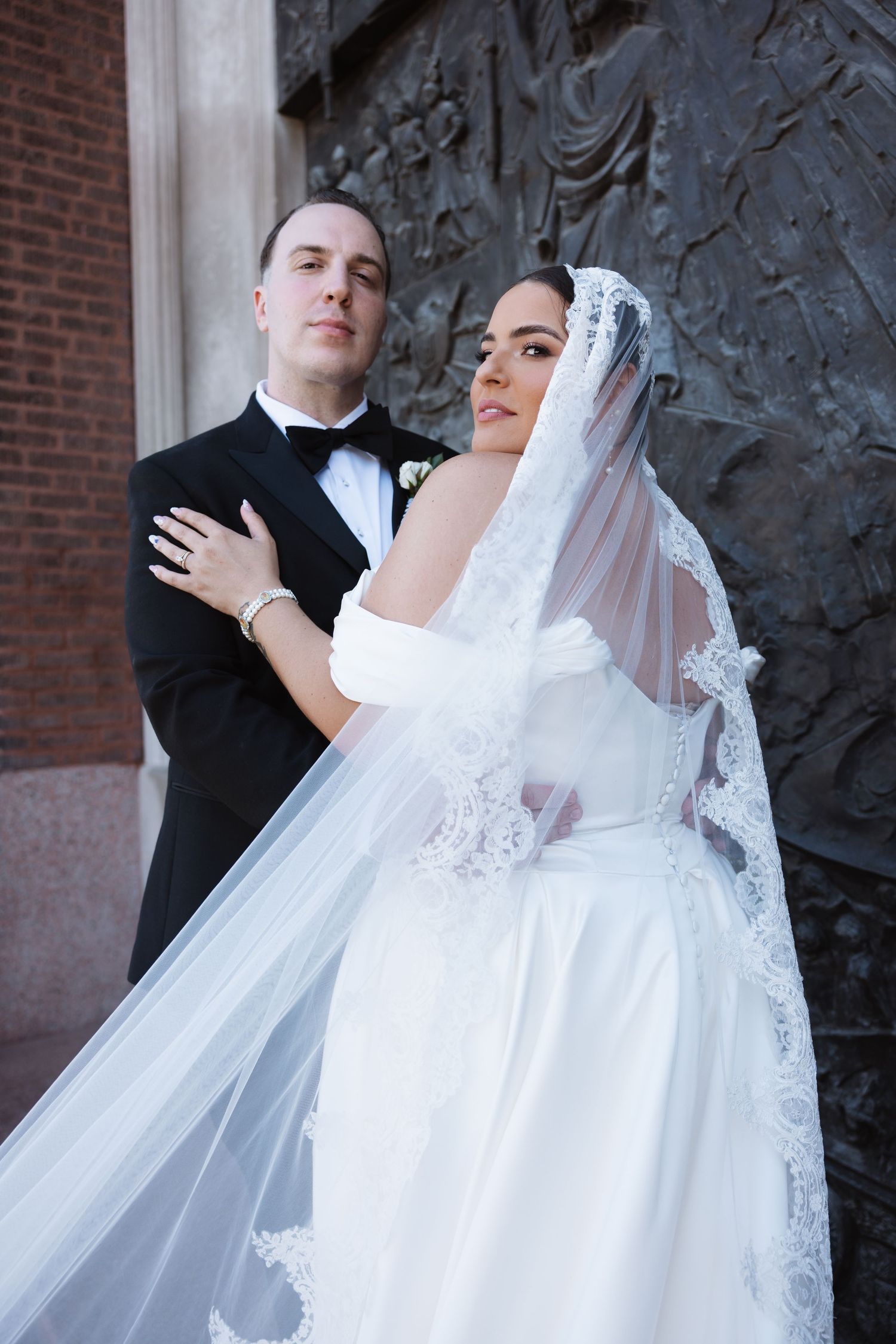 Elegant bride in lace mantilla veil and groom in black tuxedo posing together outside historic building.