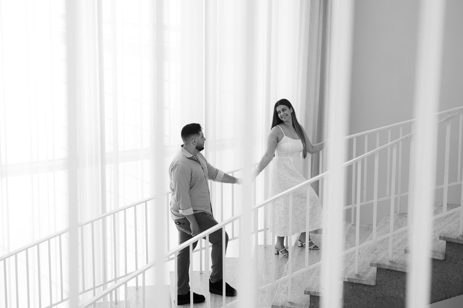 Black and white photo of a couple holding hands on elegant stairs, woman in white dress, romantic engagement shoot.