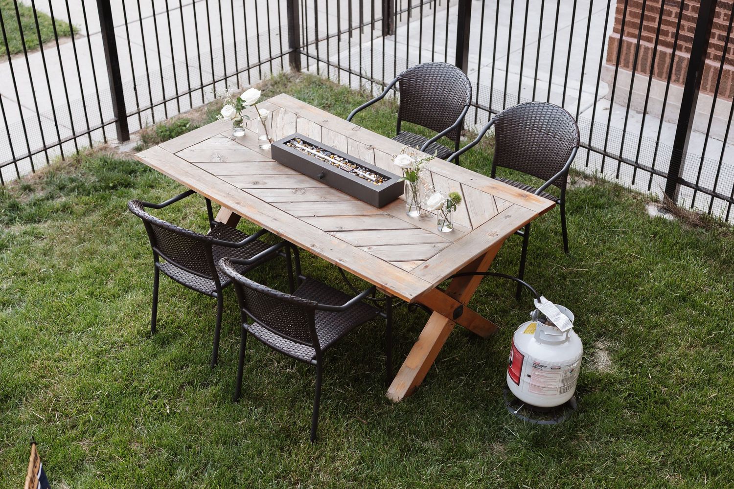 Outdoor patio dining set with wooden table, wicker chairs, and a small fire pit centerpiece.