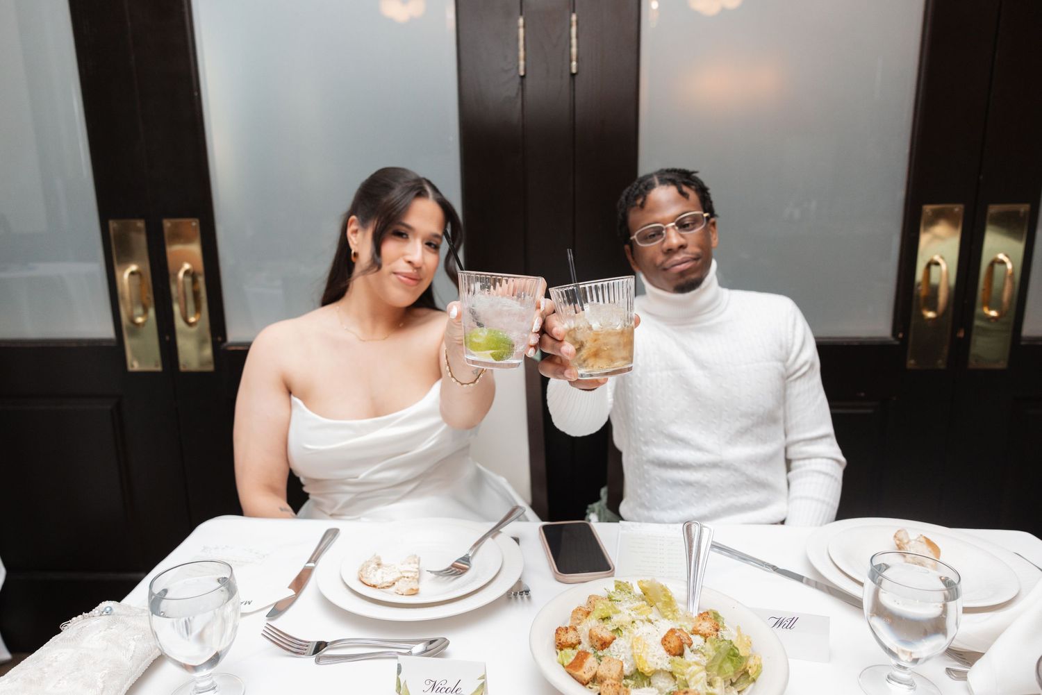 Bride and groom in white attire toasting drinks at elegant wedding dinner table.
