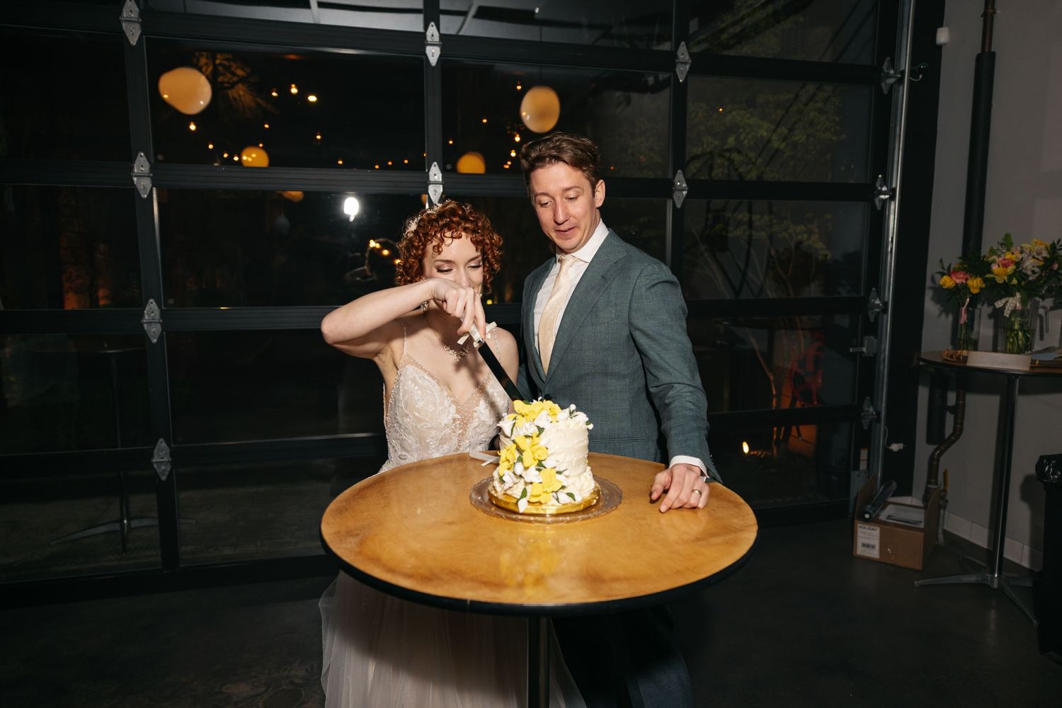 Bride and groom cut yellow floral wedding cake together at round table during evening reception.